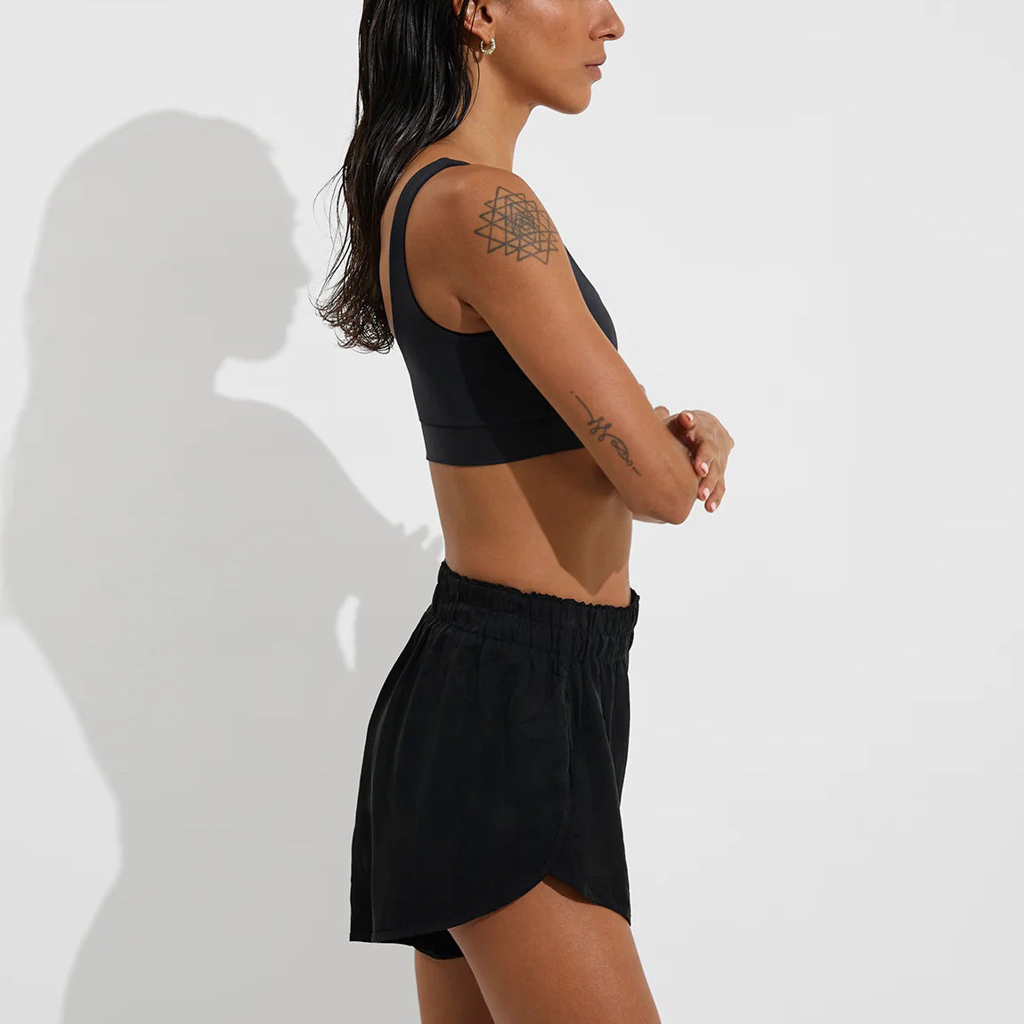 A woman stands in profile with arms crossed, wearing Left On Friday's Day Trip Short—high-waisted black athletic shorts with pockets—and a black sport liner sports bra, showcasing a geometric tattoo on her upper arm.