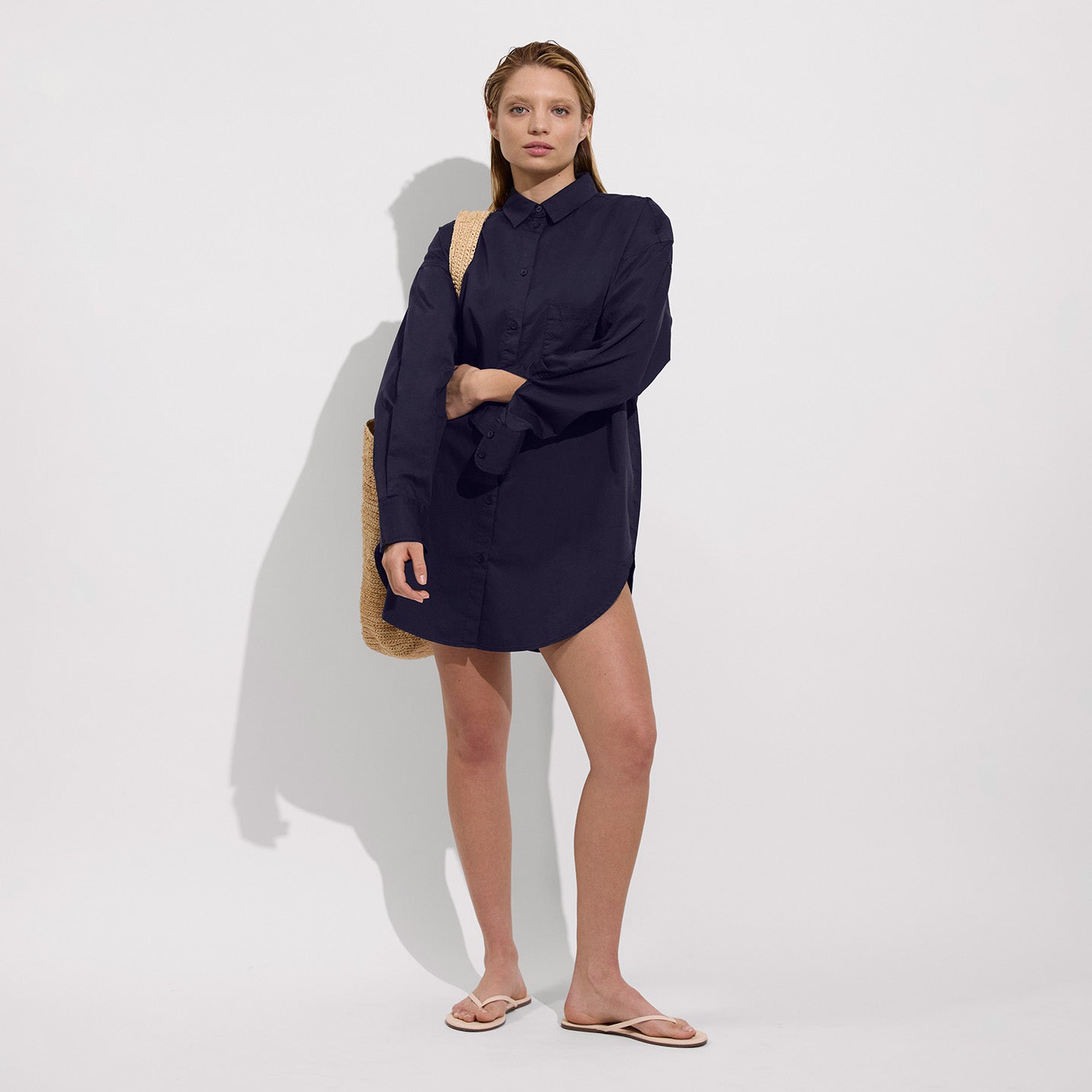 A woman stands against a plain background wearing the Left On Friday Port Shirt Dress, which has a loose fit, with flip-flops and a woven shoulder bag.