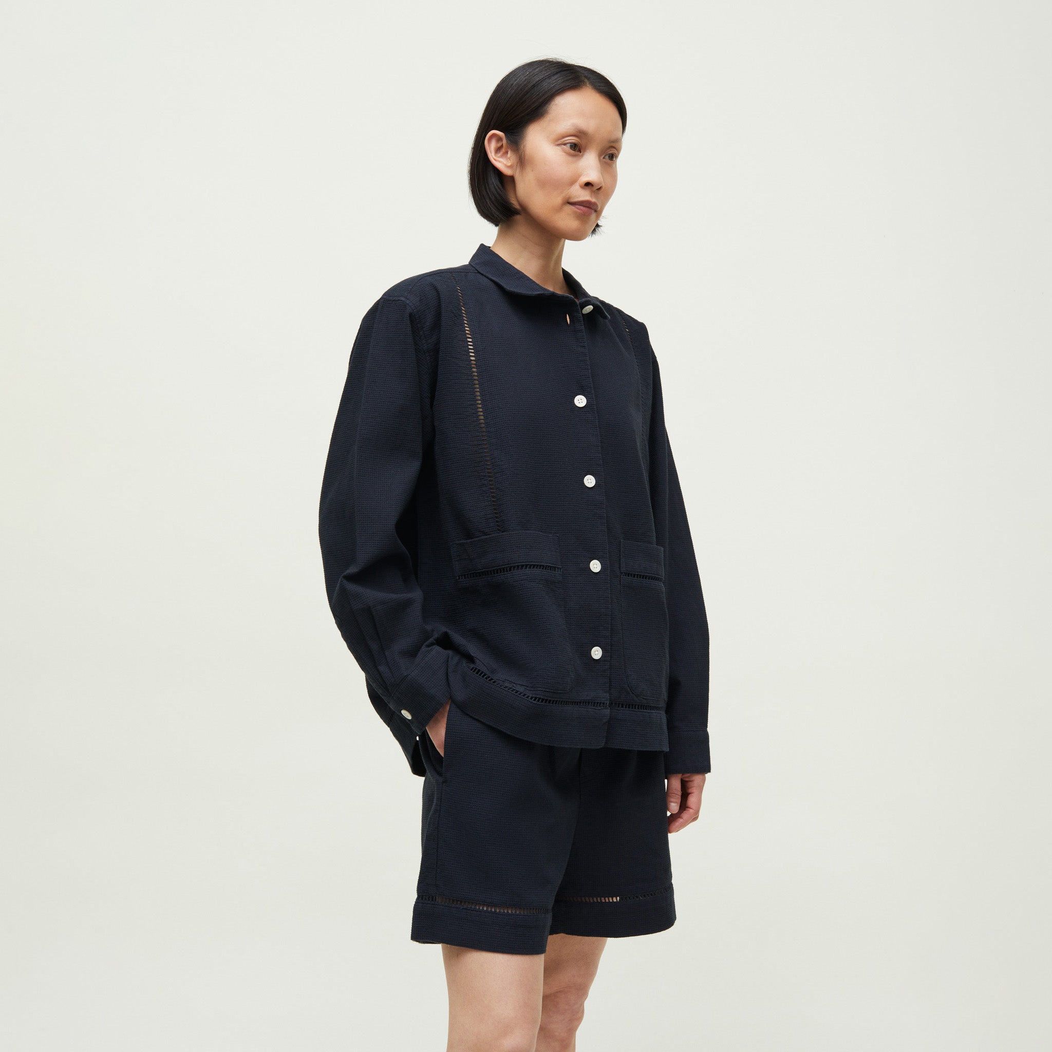 A person with short black hair wears the Aiayu Lucie Chetna Cotton Shirt Waffle in black, paired with matching black shorts, standing against a plain light background.