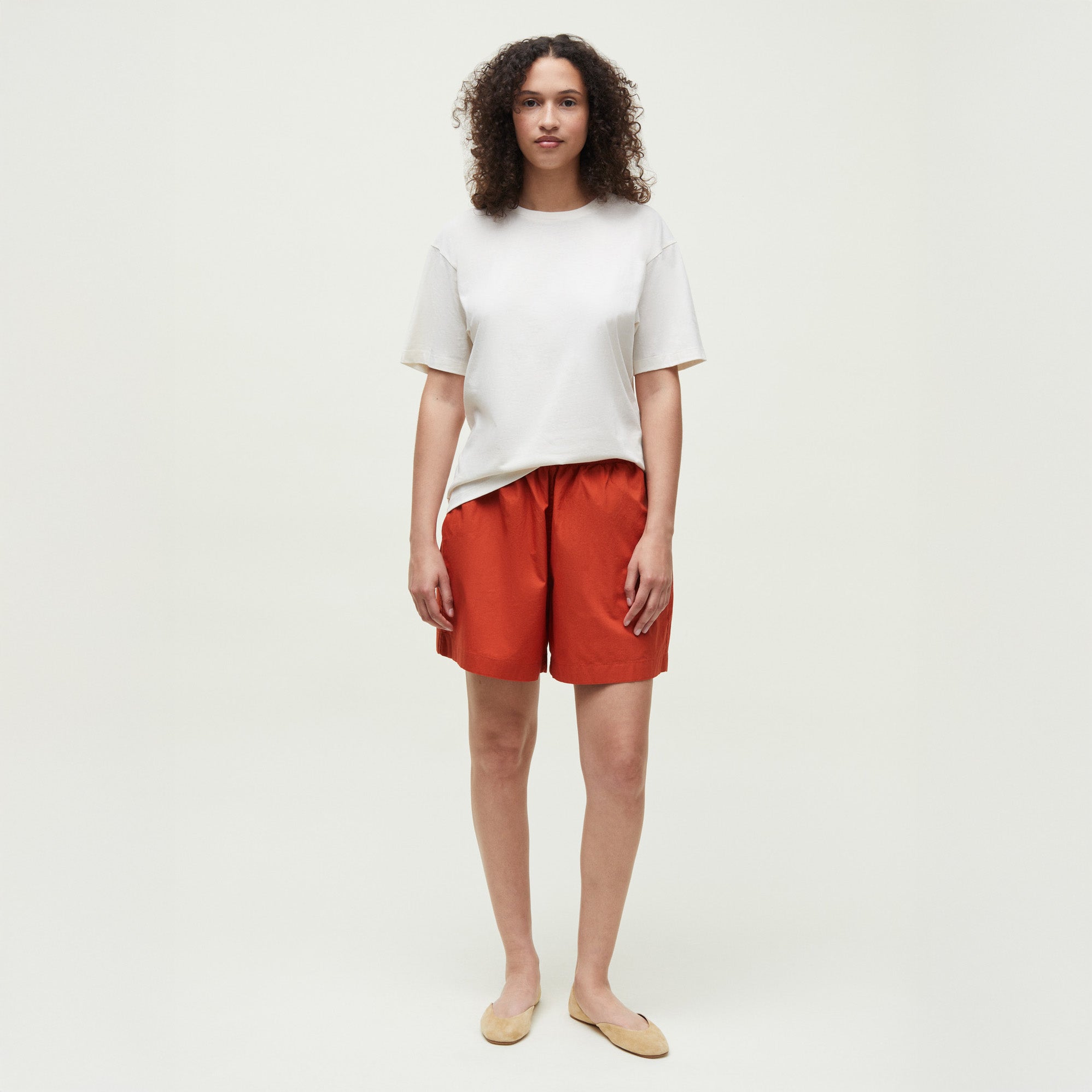 A person stands against a plain background wearing Aiayu's Lulu Poplin II white t-shirt, red organic cotton shorts with an elastic waistband, and beige flats, looking straight at the camera.