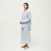 Against a plain background, a person wears an oversized Aiayu Lynette Chetna Cotton Shirt Verano in blue and white stripes, paired with a matching long skirt and beige flat shoes.