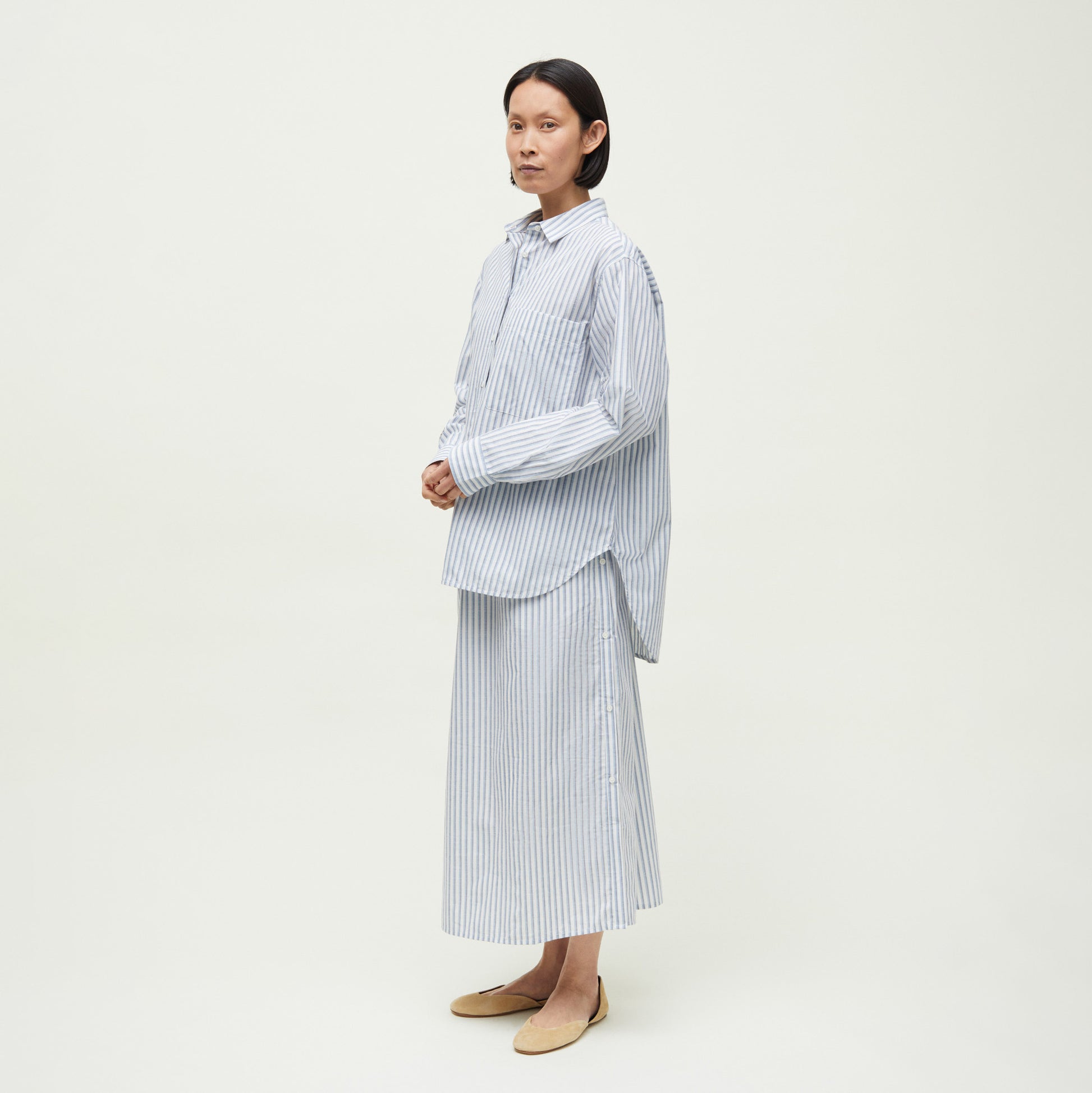 Against a plain background, a person wears an oversized Aiayu Lynette Chetna Cotton Shirt Verano in blue and white stripes, paired with a matching long skirt and beige flat shoes.