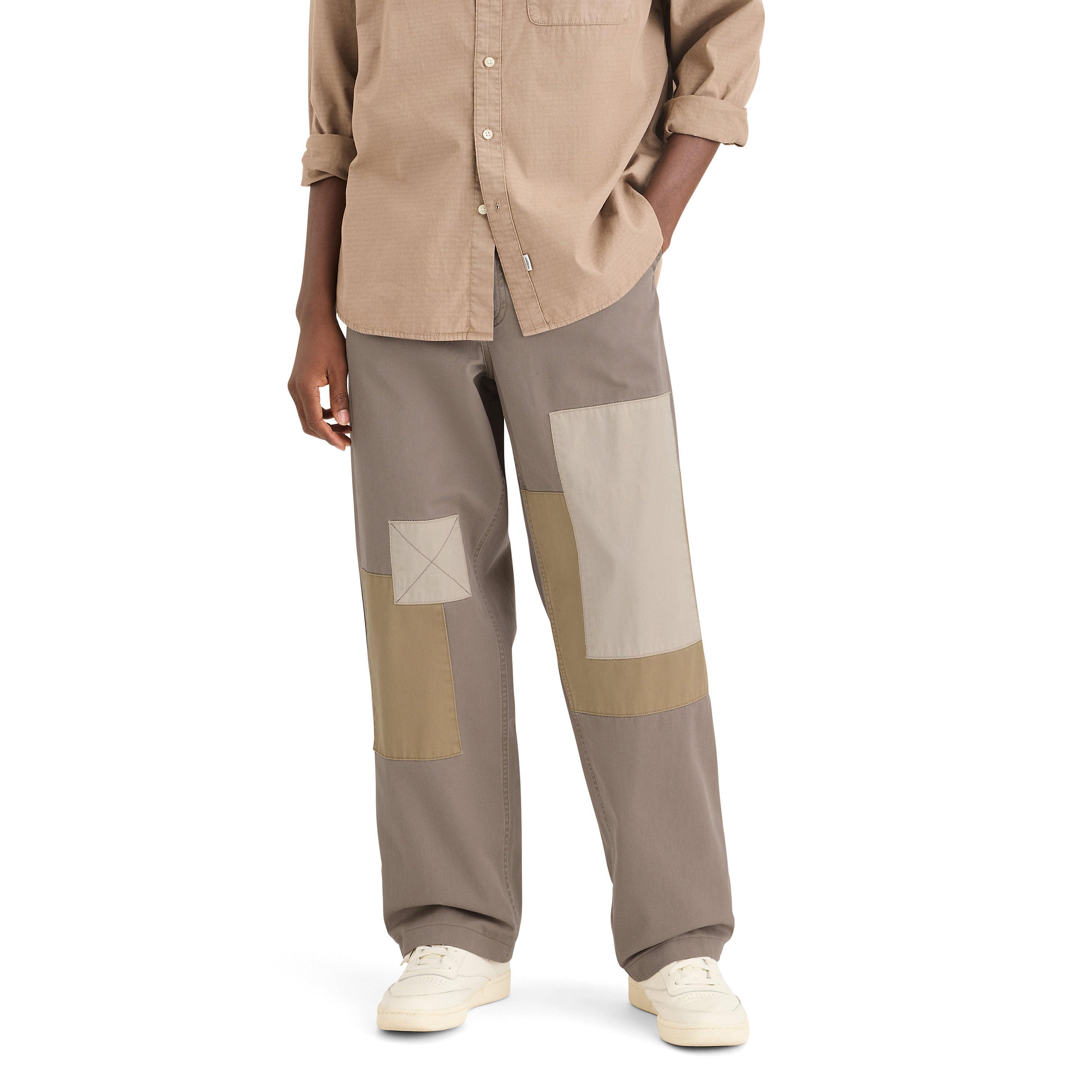 A person wearing Dockers' Genderless 86 Alpha Chino Patchwork, a beige shirt made from recycled cotton, and white sneakers stands against a plain white background.