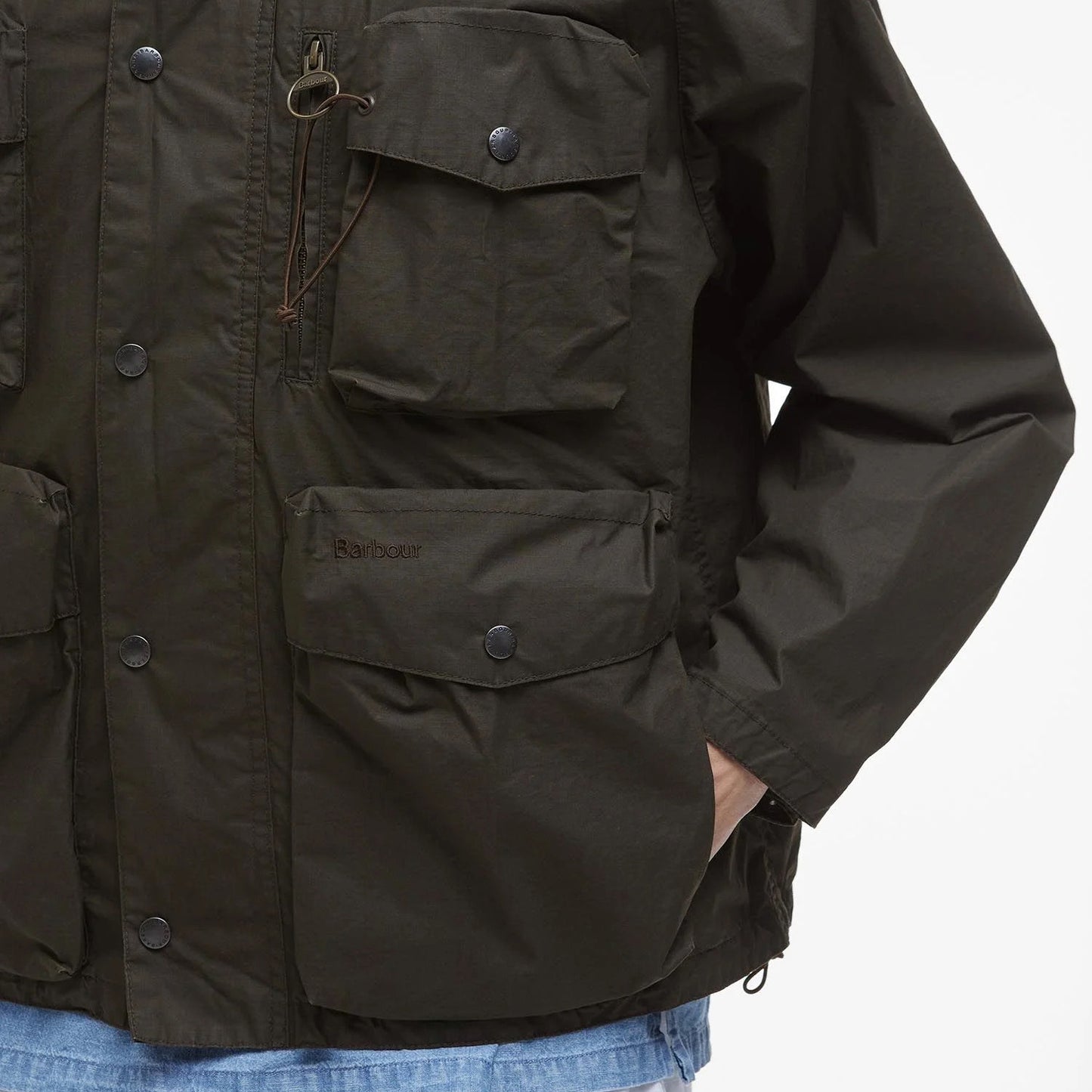 A person wears a Barbour Westmorland Showerproof Jacket, dark green, inspired by countryside living with functional details like multiple pockets. Paired with blue jeans, this ensemble features drawstring and button accents.