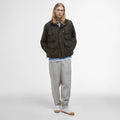 A person with long hair stands against a plain white background, wearing a Barbour Westmorland Showerproof Jacket over a light blue shirt and light pants, combining functionality with a touch of countryside style.