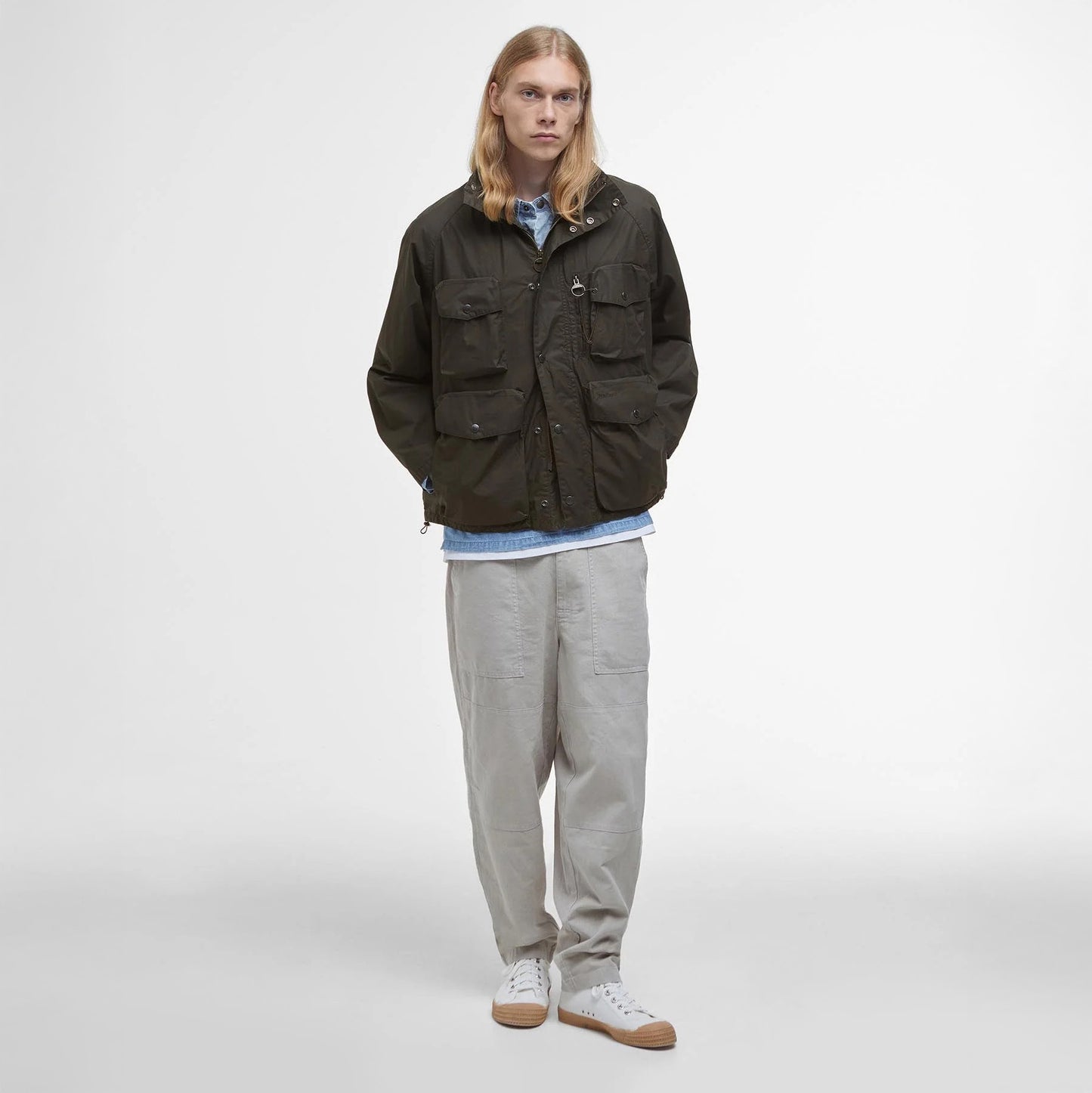A person with long hair stands against a plain white background, wearing a Barbour Westmorland Showerproof Jacket over a light blue shirt and light pants, combining functionality with a touch of countryside style.
