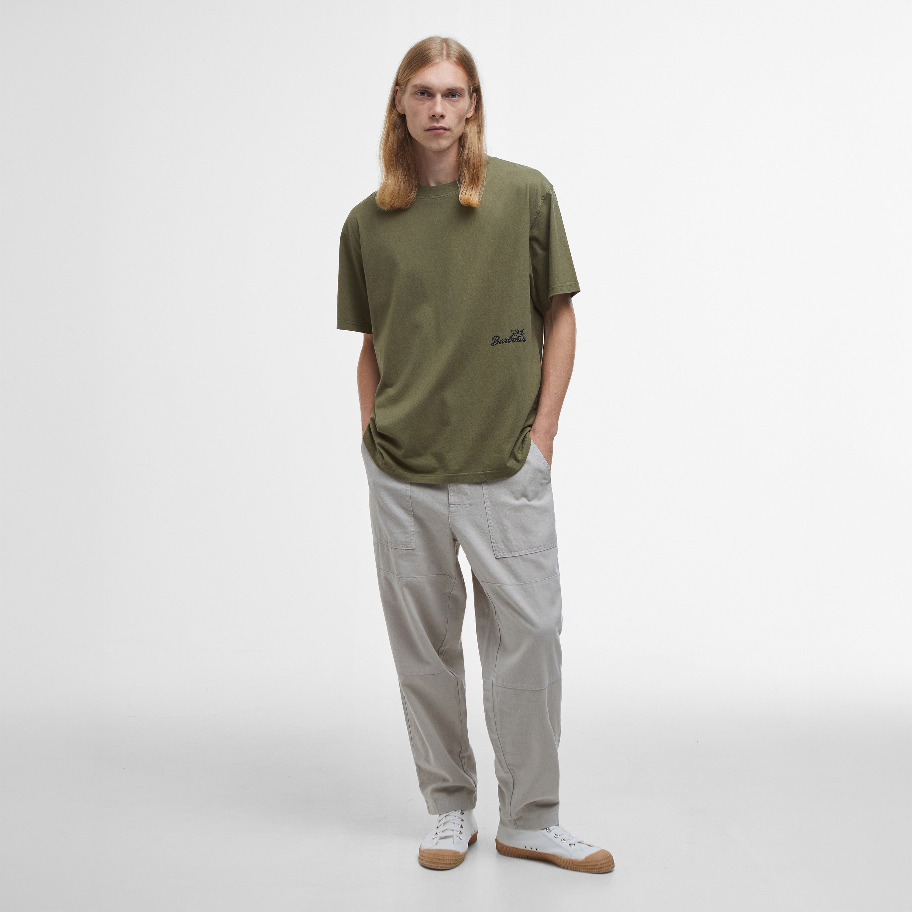 A person with long blonde hair stands wearing an oversized Regence Graphic T-Shirt by Barbour, light gray pants, and white sneakers against a plain background.