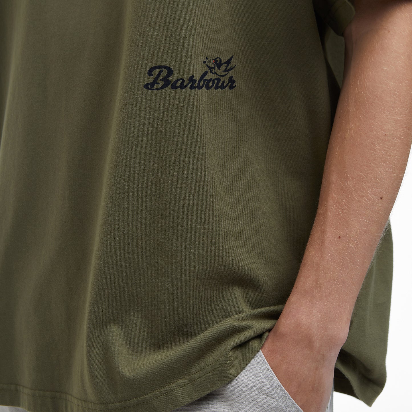 A close-up features someone in an archive-inspired olive green Barbour Regence Graphic T-Shirt and light gray pants, with their hand in the pocket. The partial view of the torso and arm emphasizes the understated elegance of classic design.