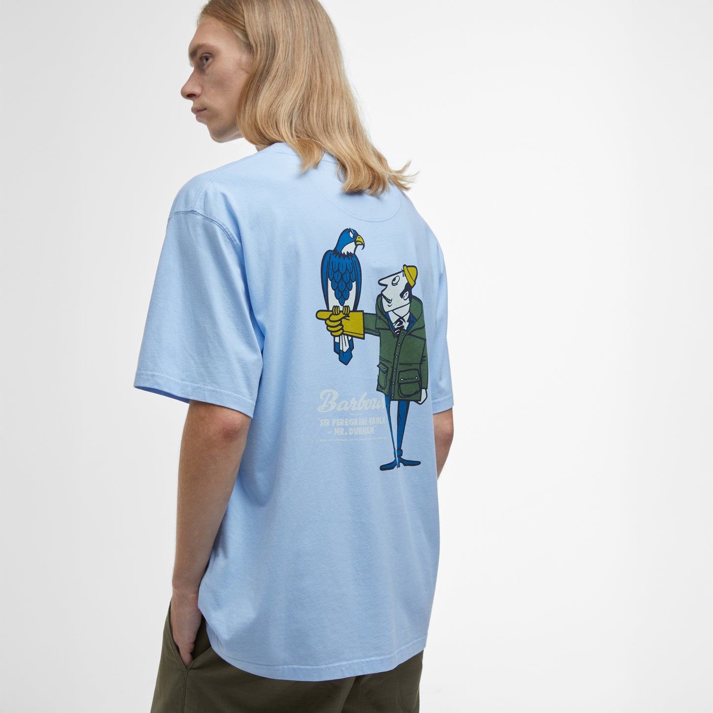 Wearing Barbour's Newclyde Graphic T-Shirt, an oversized, archive-inspired light blue tee featuring a back graphic of a cartoon character with a bird, standing against a plain background.