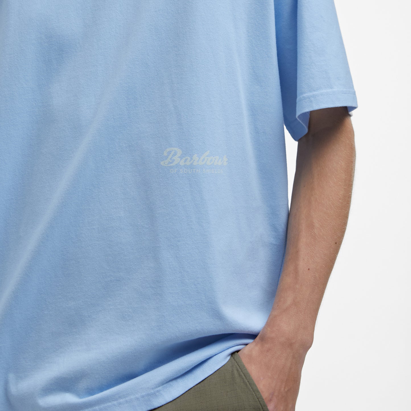 A person wearing a light blue Barbour Newclyde Graphic T-Shirt with one hand in pocket against a plain white background.