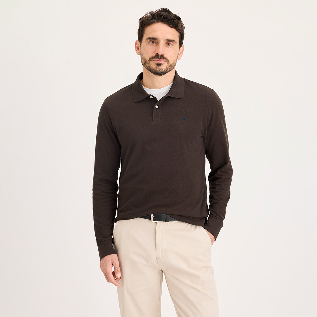 A man stands against a plain background, dressed in the Dockers Ls Polo, Mole long-sleeve shirt paired with light-colored trousers and a belt.