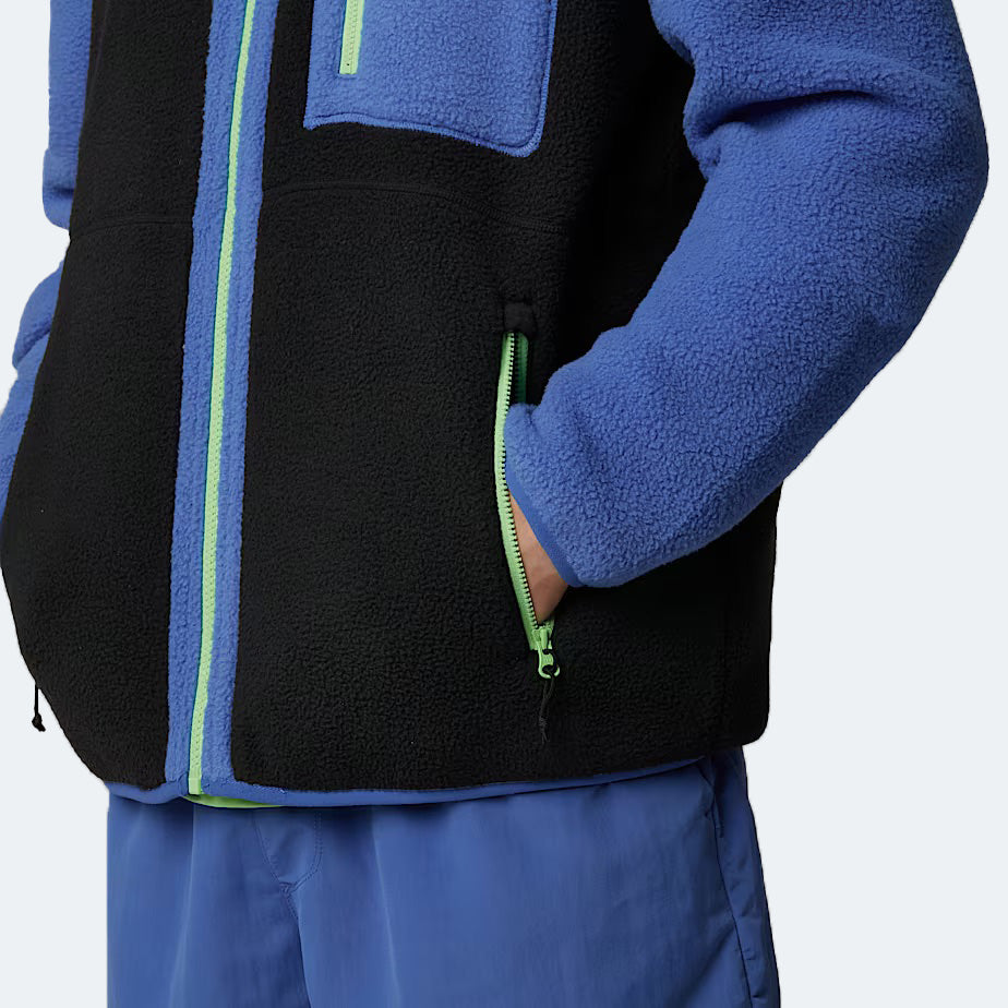 A person wearing The North Face M Yumiori Full Zip, a blue and black midweight fleece jacket with green zippers, paired with blue pants, casually places their hand in the jacket pocket.
