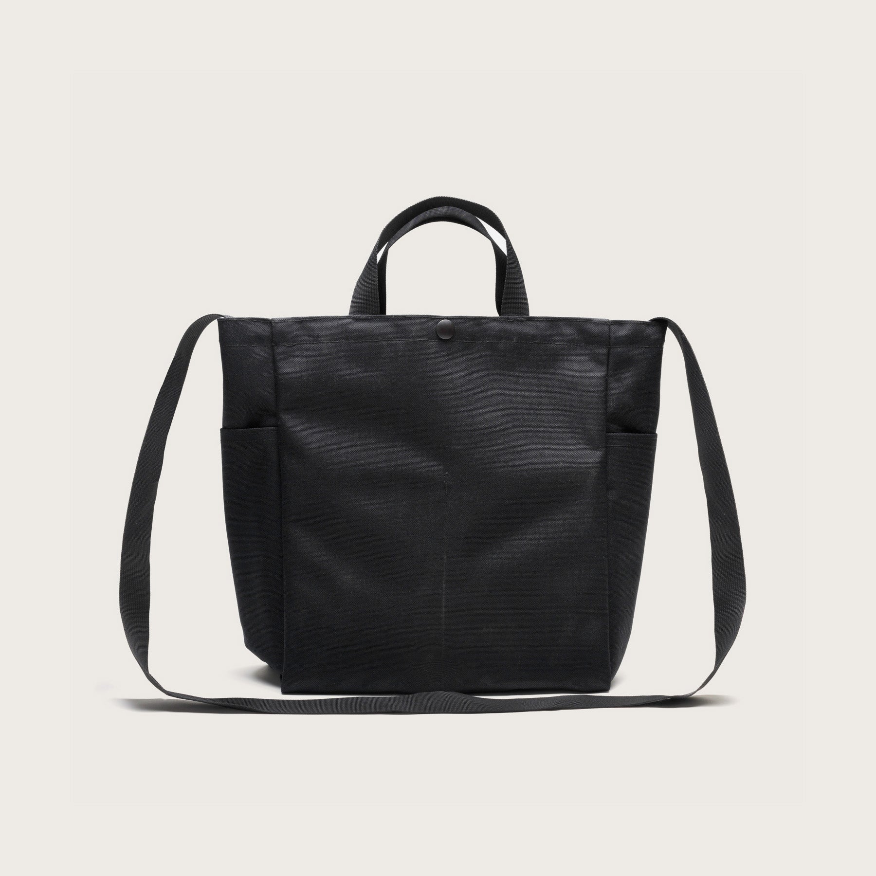 The Multi Pocket Bag Medium by Bags in Progress is a black nylon canvas tote with short handles, a long adjustable shoulder strap, water-repellent finish, and side pockets, shown against a plain white background.