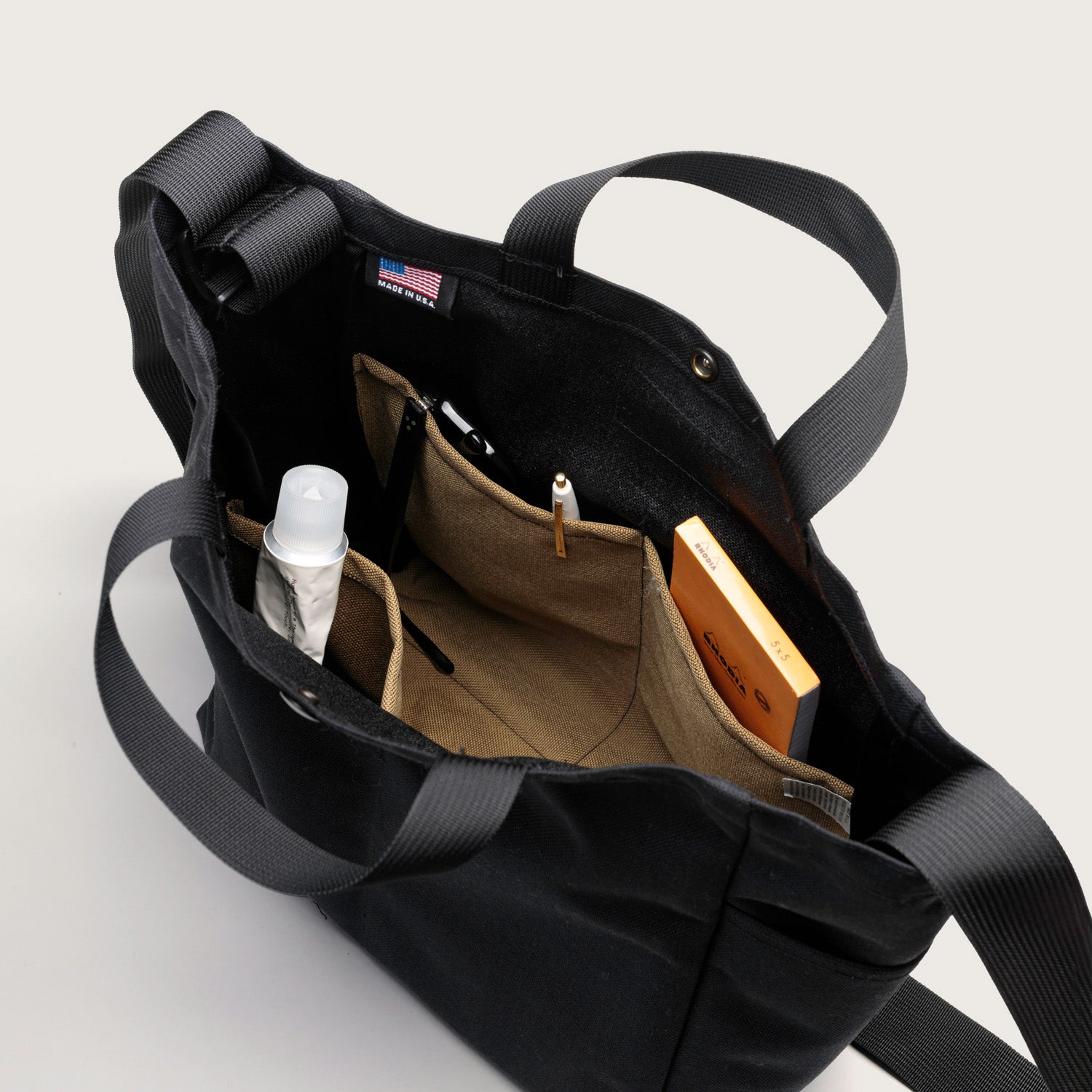 The Multi Pocket Bag Medium by Bags in Progress, shown open in black nylon canvas, features a beige water-repellent interior with pockets holding a spray bottle, pens, and a small orange box.