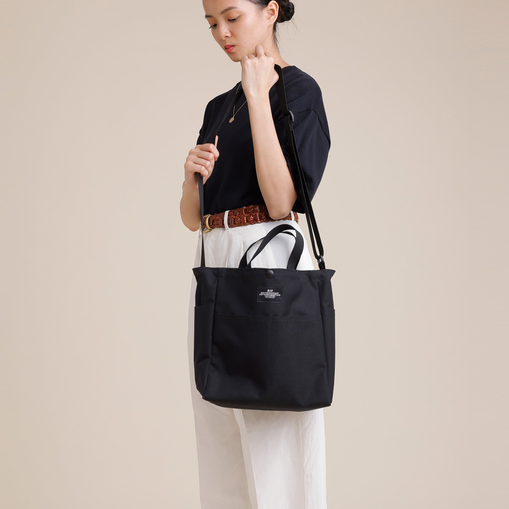 A woman in a black shirt and white pants carries the Bags in Progress Multi Pocket Bag Medium, a black nylon canvas shoulder bag with hand straps and water-repellent material, against a plain beige background.