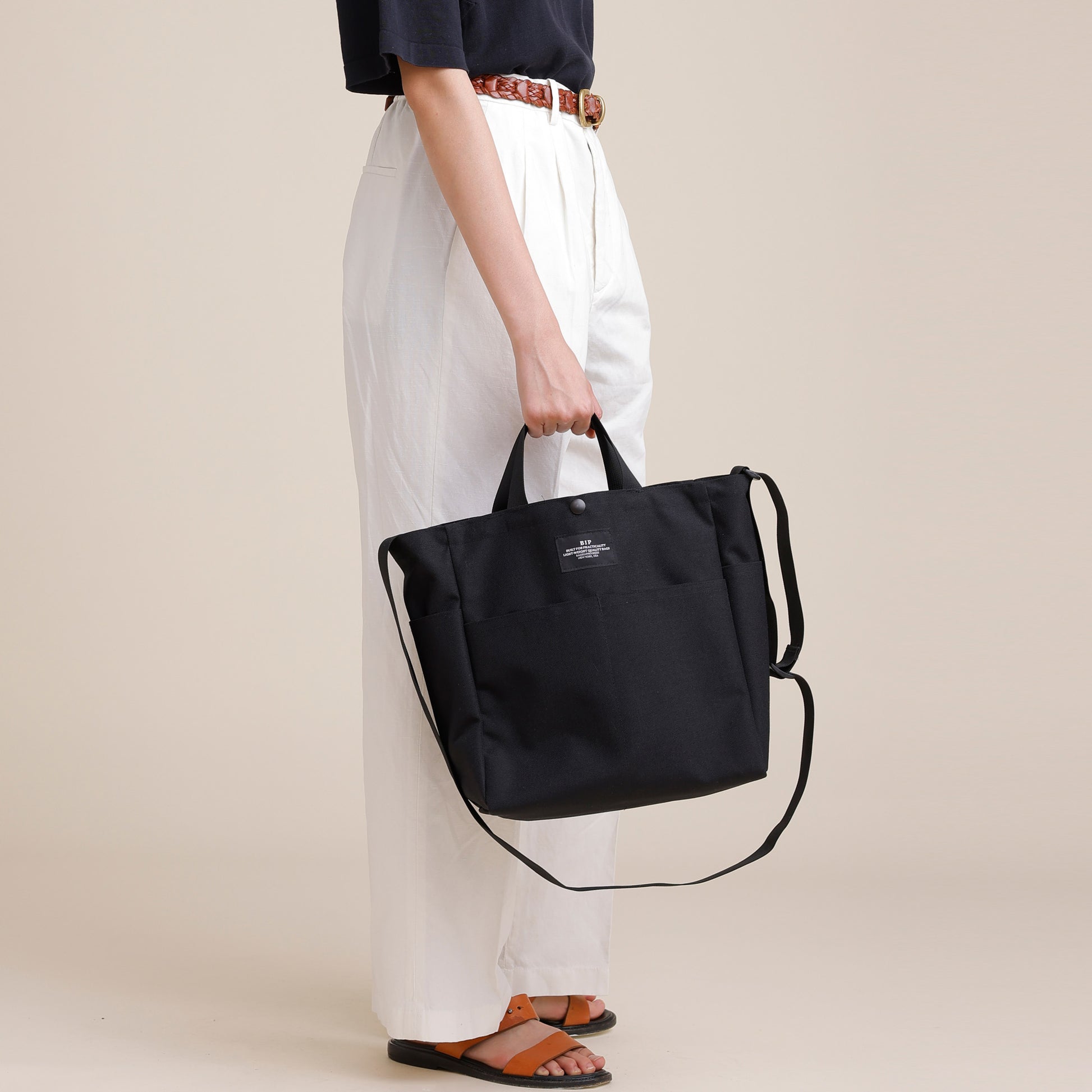 A person stands sideways in white pants, holding the Bags in Progress Multi Pocket Bag Medium—black, water-repellent nylon canvas with short handles and a shoulder strap—against a plain background.