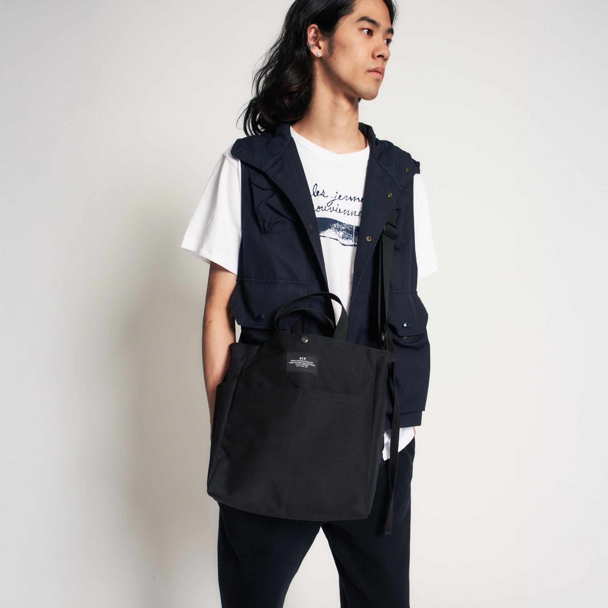A person stands against a plain background wearing a white t-shirt, dark vest, and dark pants, holding the Bags in Progress Multi Pocket Bag Medium, a large black nylon canvas shoulder bag.