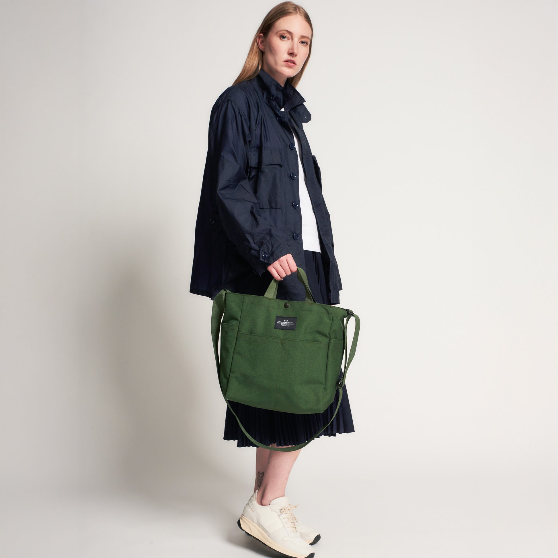 A person stands against a plain background wearing a navy jacket, white shirt, dark skirt, and white sneakers, holding a Bags in Progress Multi Pocket Bag Medium in large green nylon canvas.
