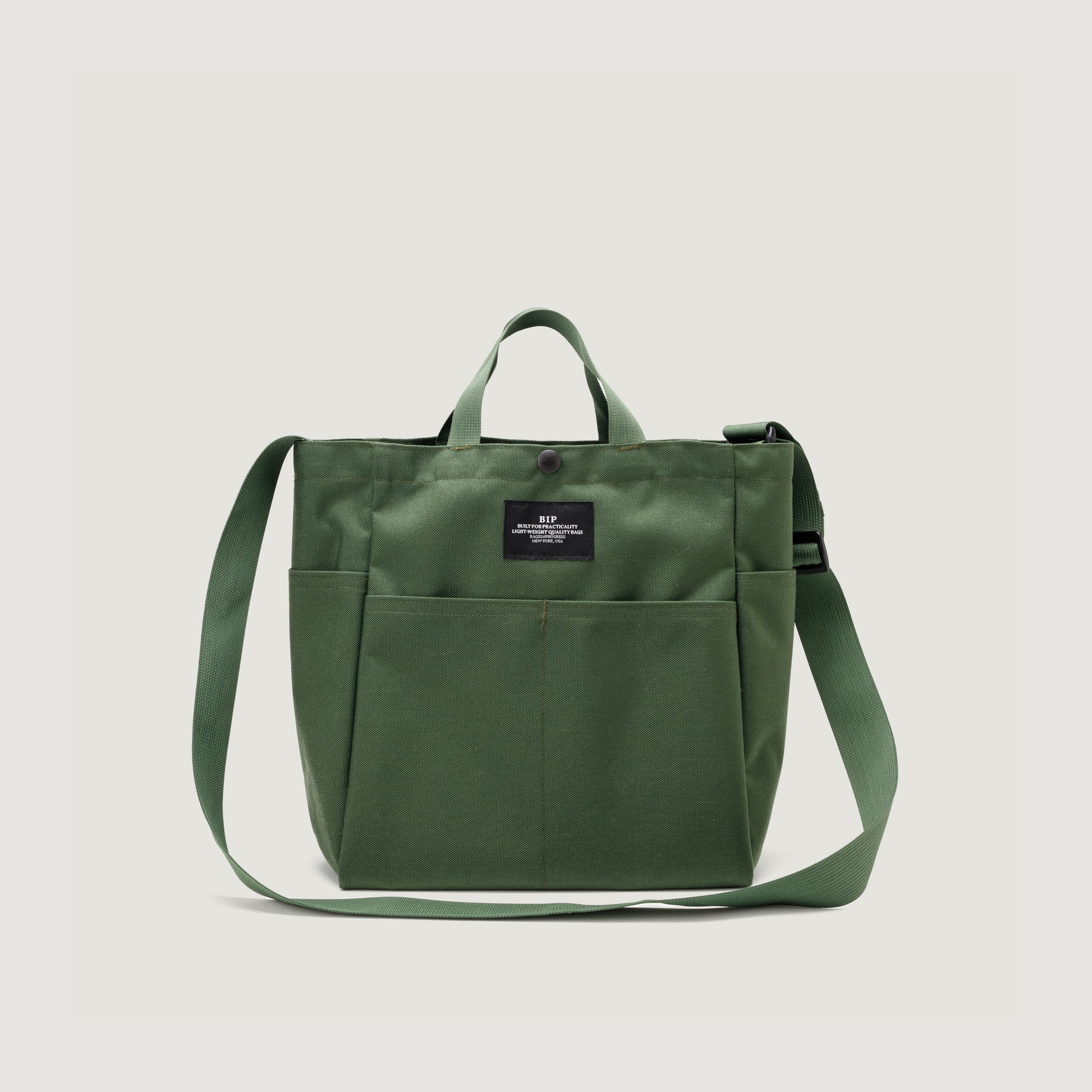 The Multi Pocket Bag Medium by Bags in Progress is crafted from water-repellent green nylon canvas with front pockets, short handles, a long adjustable strap, and a black front label.