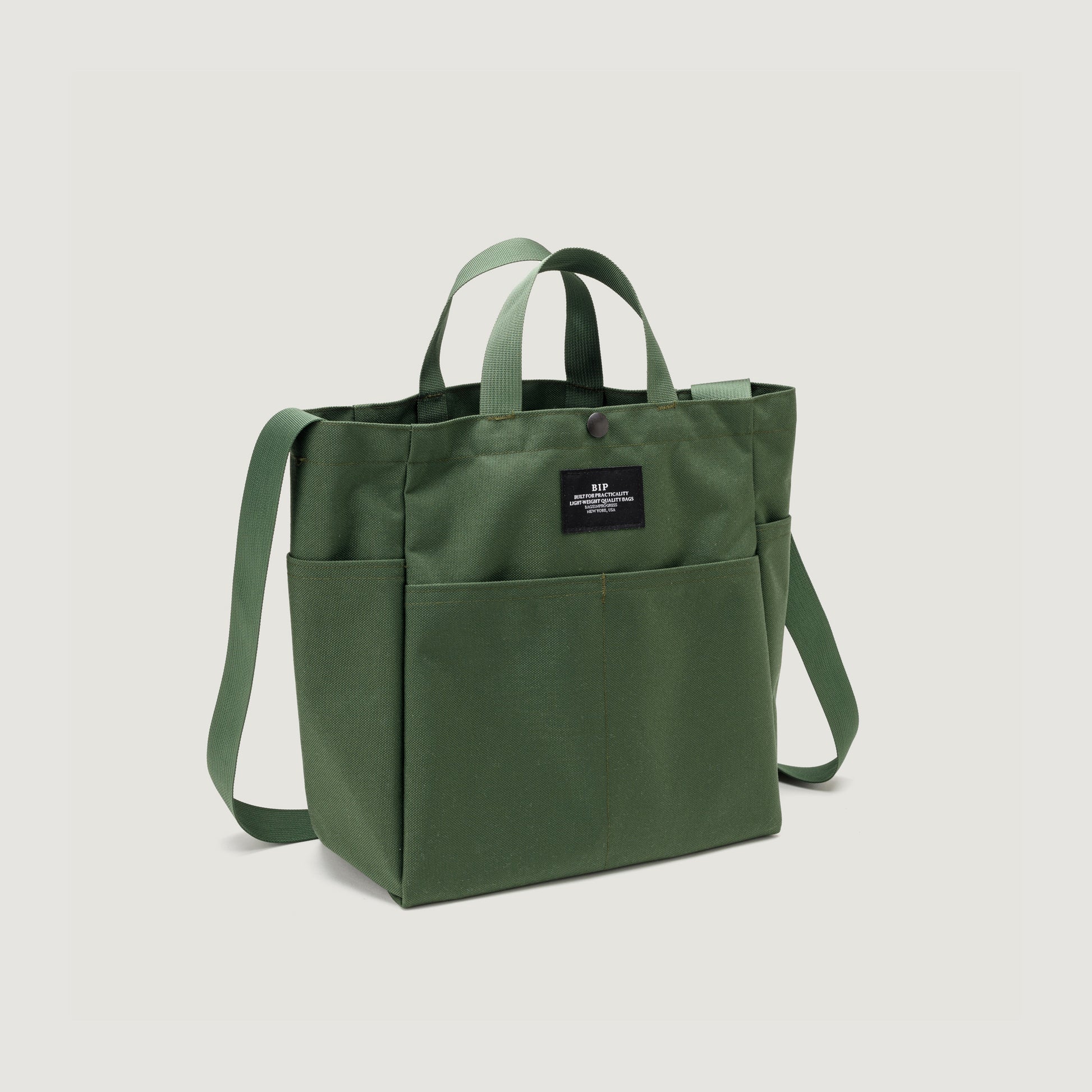 The Multi Pocket Bag Medium by Bags in Progress is a green nylon canvas tote with two handles, a shoulder strap, front pockets, water-repellent finish, and a black front label—combining style and practicality for daily use.