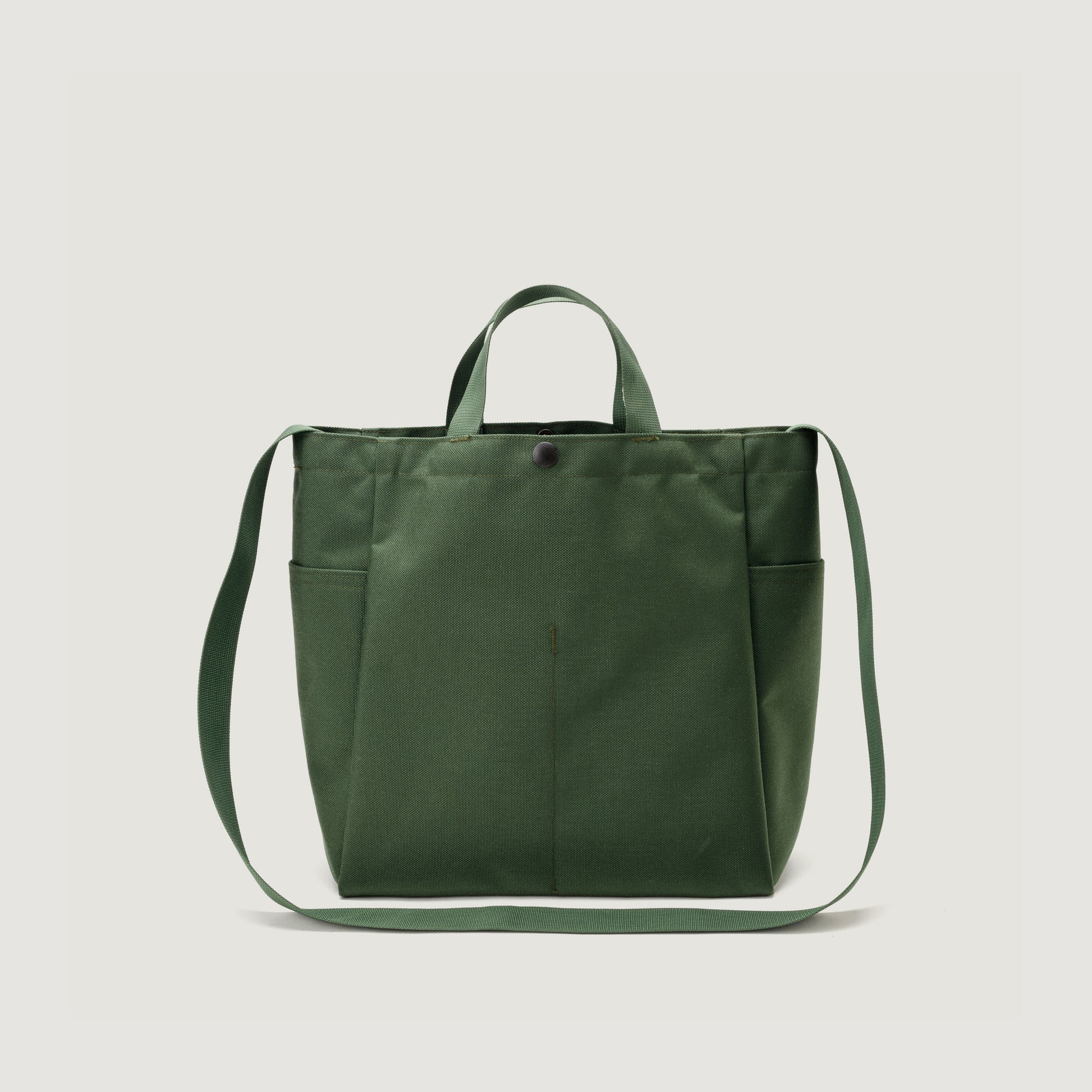 The Bags in Progress Multi Pocket Bag Medium is a green, water-repellent nylon canvas shoulder bag with two short handles, an adjustable strap, front pocket, and snap closure, shown on a plain white background.