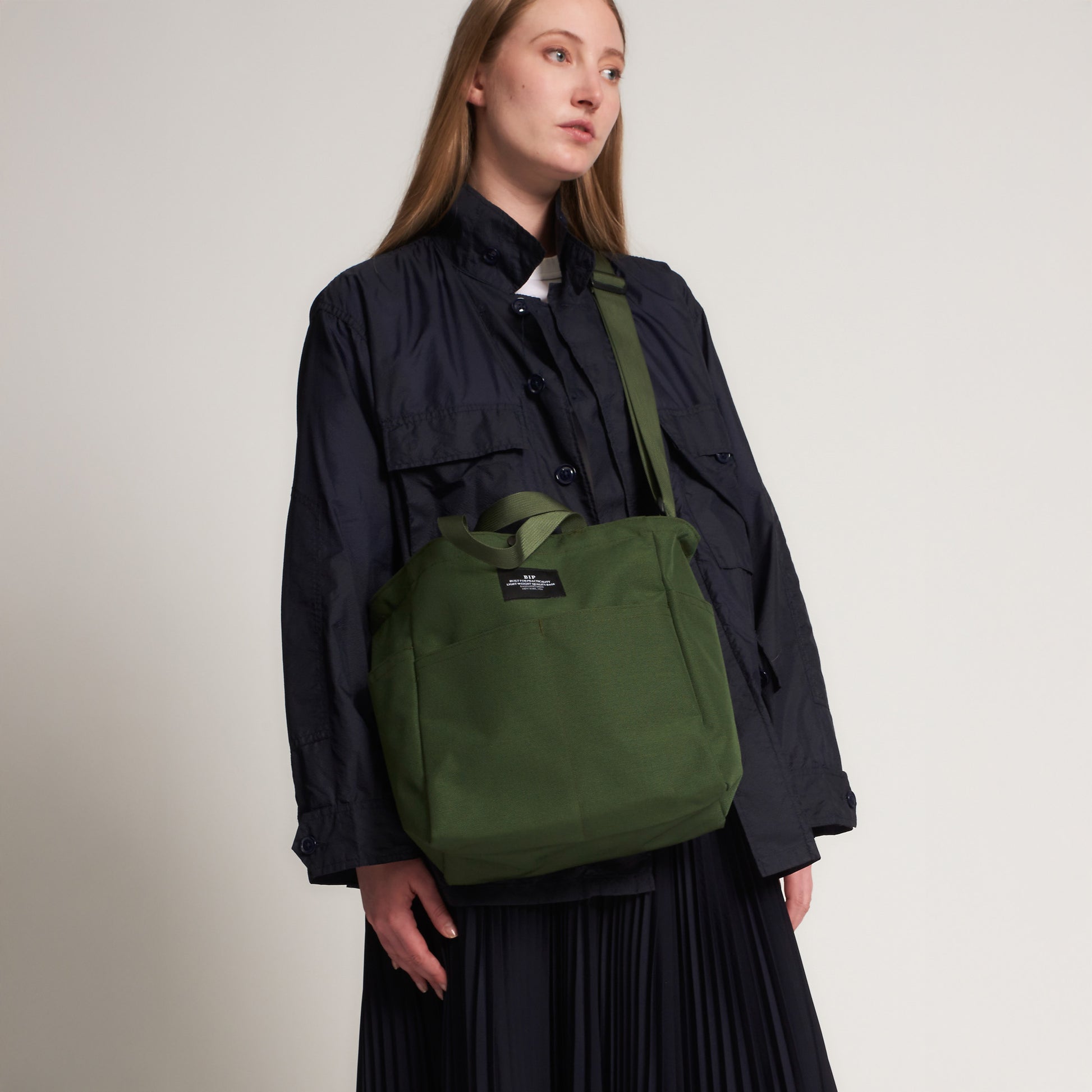 A person stands against a plain background, wearing a black buttoned jacket and pleated skirt, carrying the Bags in Progress Multi Pocket Bag Medium—a large green water-repellent nylon canvas shoulder bag.