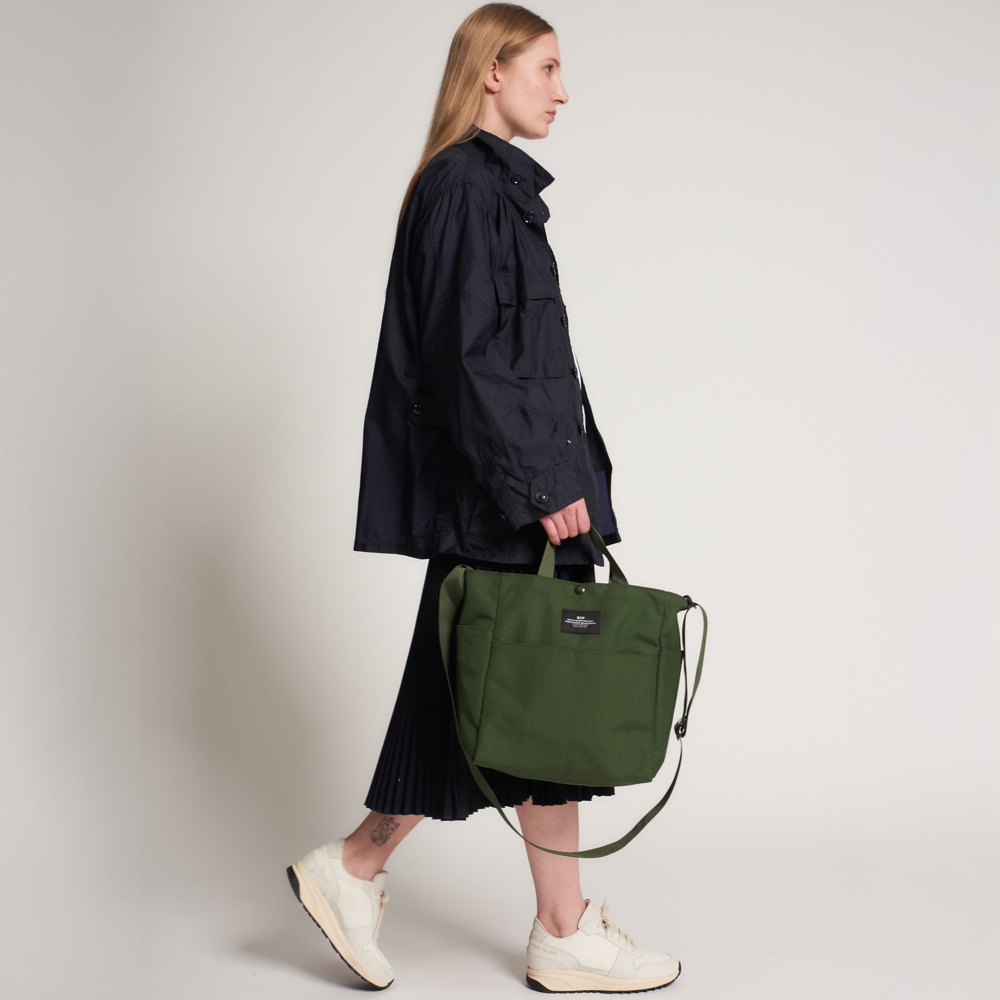 Person in a black jacket, black pleated skirt, and white sneakers carrying the Bags in Progress Multi Pocket Bag Medium—a water repellent nylon canvas shoulder bag with a green strap—standing against a plain background.