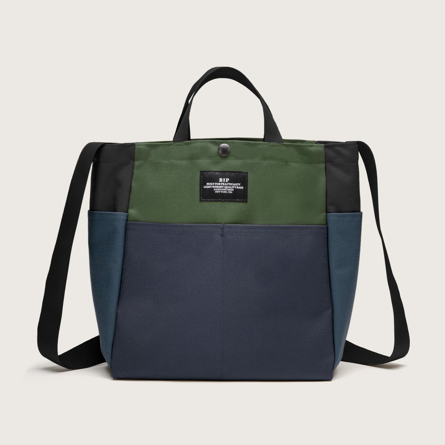 The Multi Pocket Bag Medium by Bags in Progress is a water-repellent nylon canvas tote in green, navy, and black with two handles, a shoulder strap, front label patch, and multiple pockets.