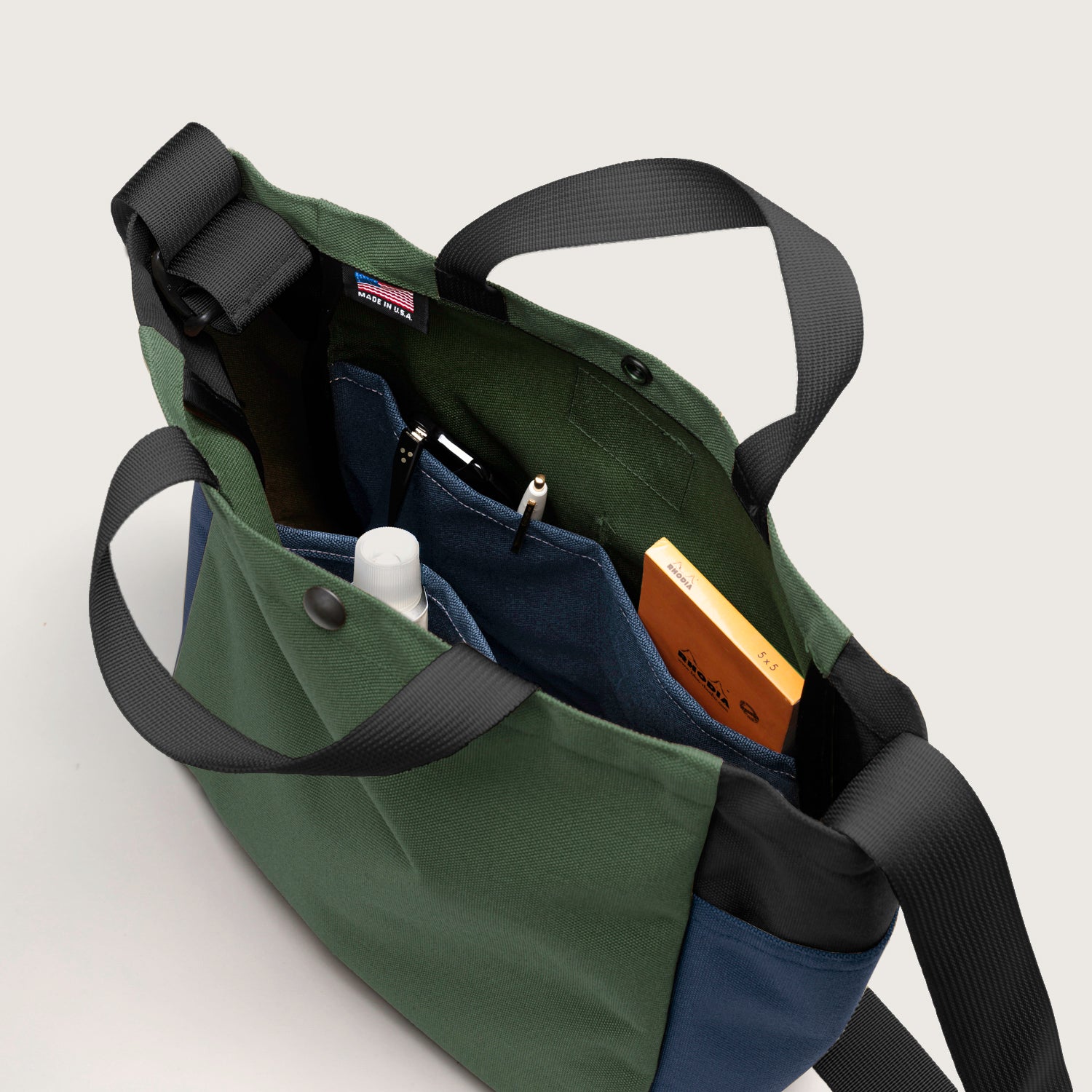The Multi Pocket Bag Medium by Bags in Progress features black straps and is made from water repellent nylon canvas in green, blue, and black, with interior pockets to hold items like a white bottle, notebook, and yellow box.