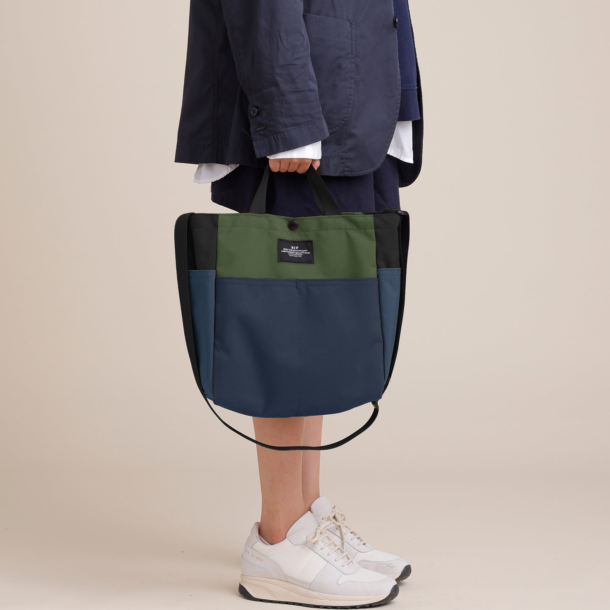 A person in a navy jacket and white sneakers holds the Bags in Progress Multi Pocket Bag Medium, a color-blocked shoulder bag made from water-repellent nylon canvas in green, blue, and black, against a plain background.