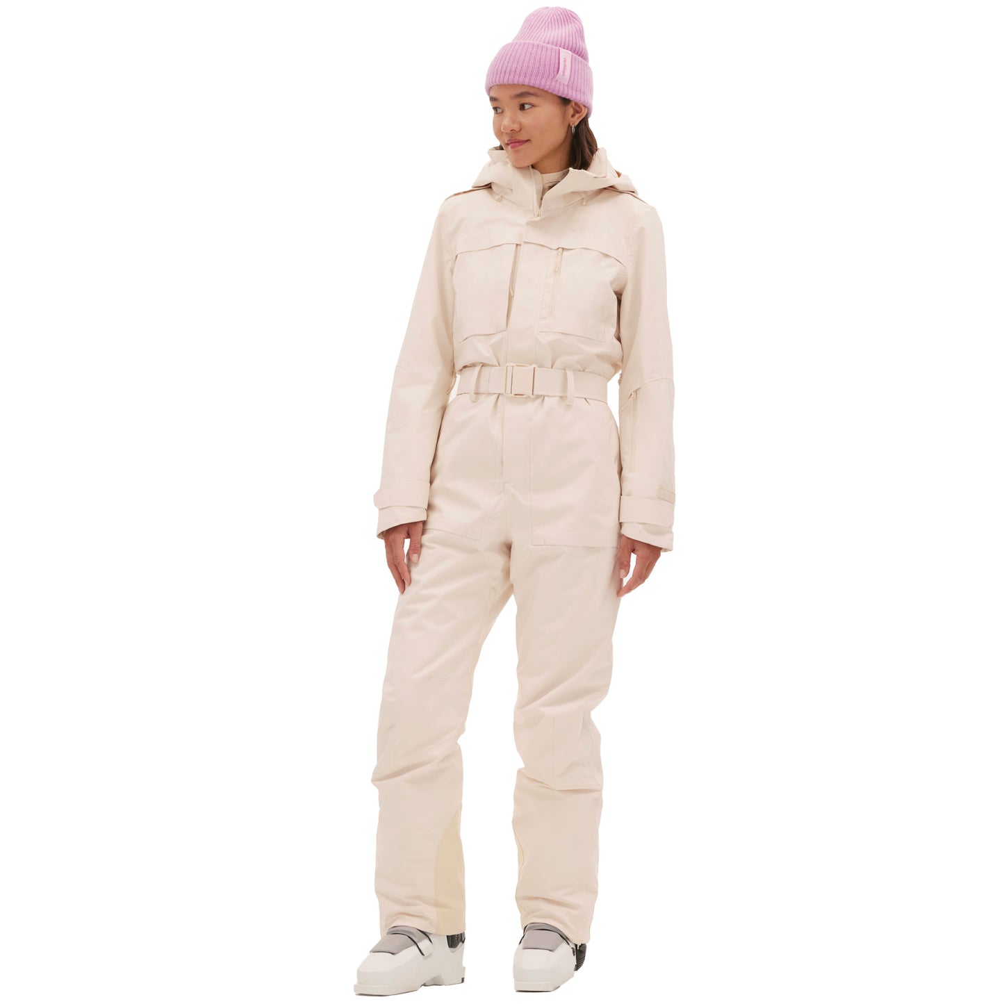 Wearing the vintage racing-style Murphy Ski Suit by Halfdays in light pink, accessorized with a matching beanie and white boots, a person stands gracefully while gazing to the side.