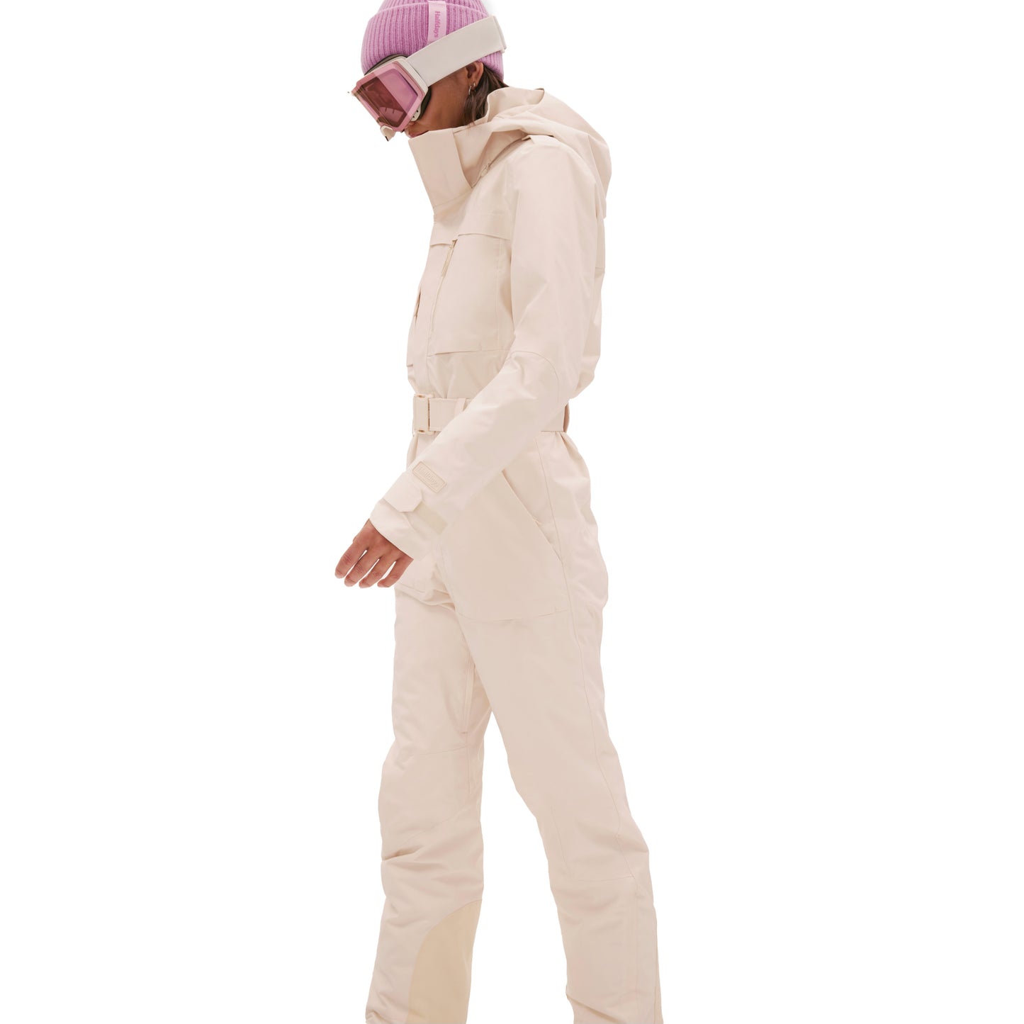A person dressed in the cream-colored Murphy Ski Suit by Halfdays, accessorized with a pink beanie and ski goggles, is viewed in profile against a white backdrop.