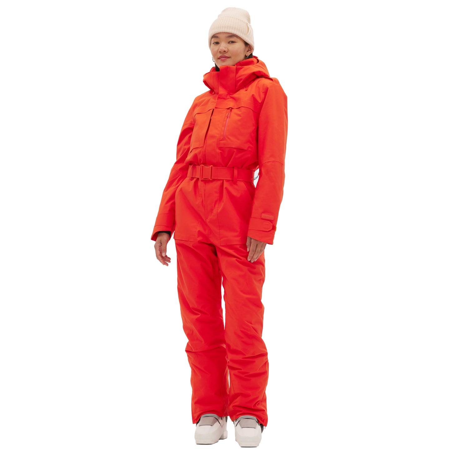 A person is wearing the Murphy Ski Suit by Halfdays, an insulated, bright orange, one-piece with a vintage racing-style design, paired with a white beanie and matching shoes against a plain white background.