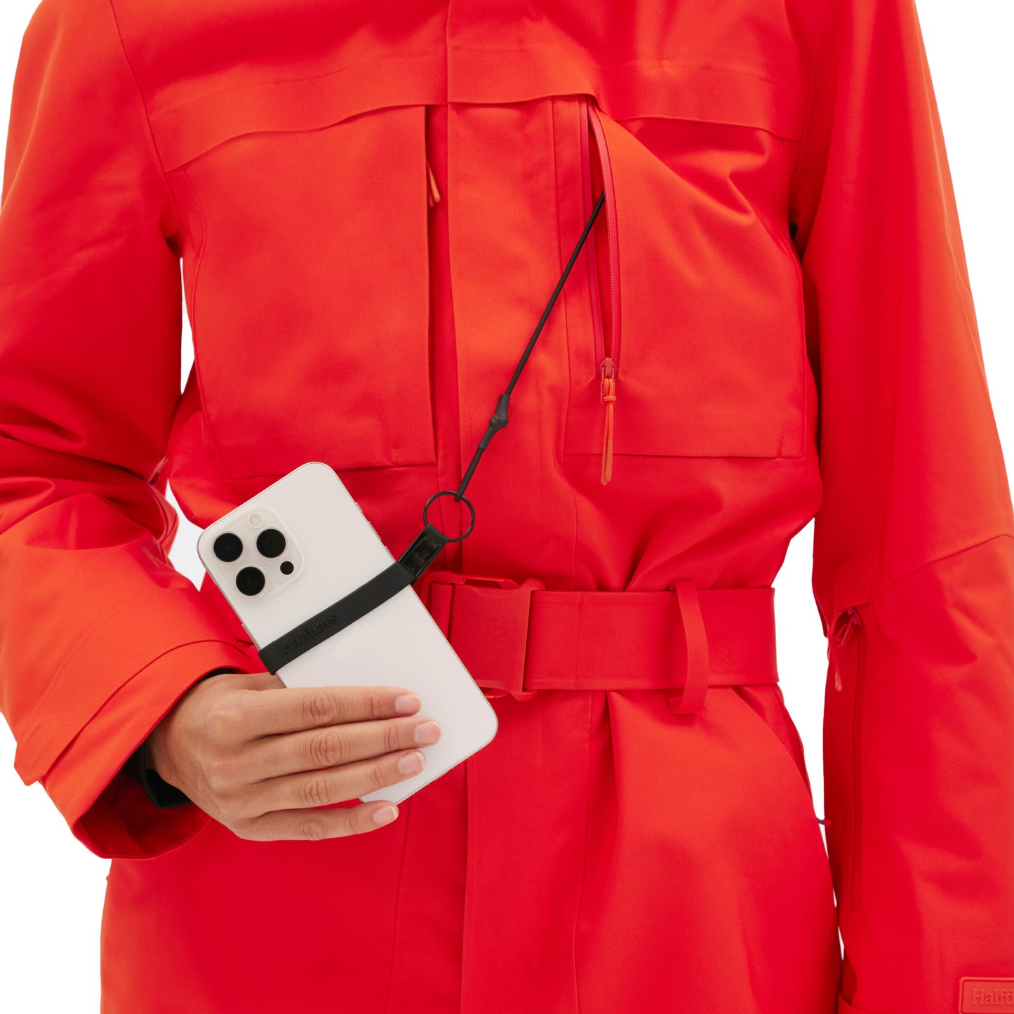 A person in a Murphy Ski Suit by Halfdays, featuring an orange jacket in a vintage racing style, is holding a smartphone with a black strap attached.