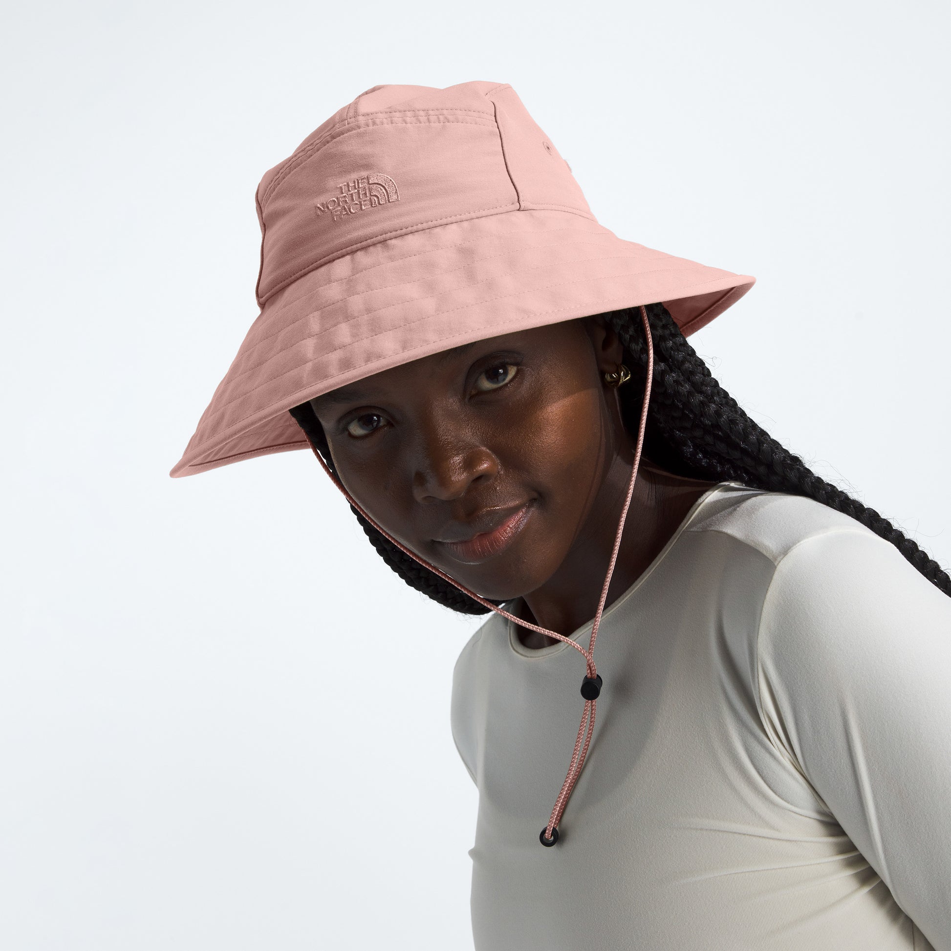 A person wearing The North Face's W Class V Brimmer, a pink wide-brimmed hat with a chin cord, paired with a beige top for sun protection, looks towards the camera.