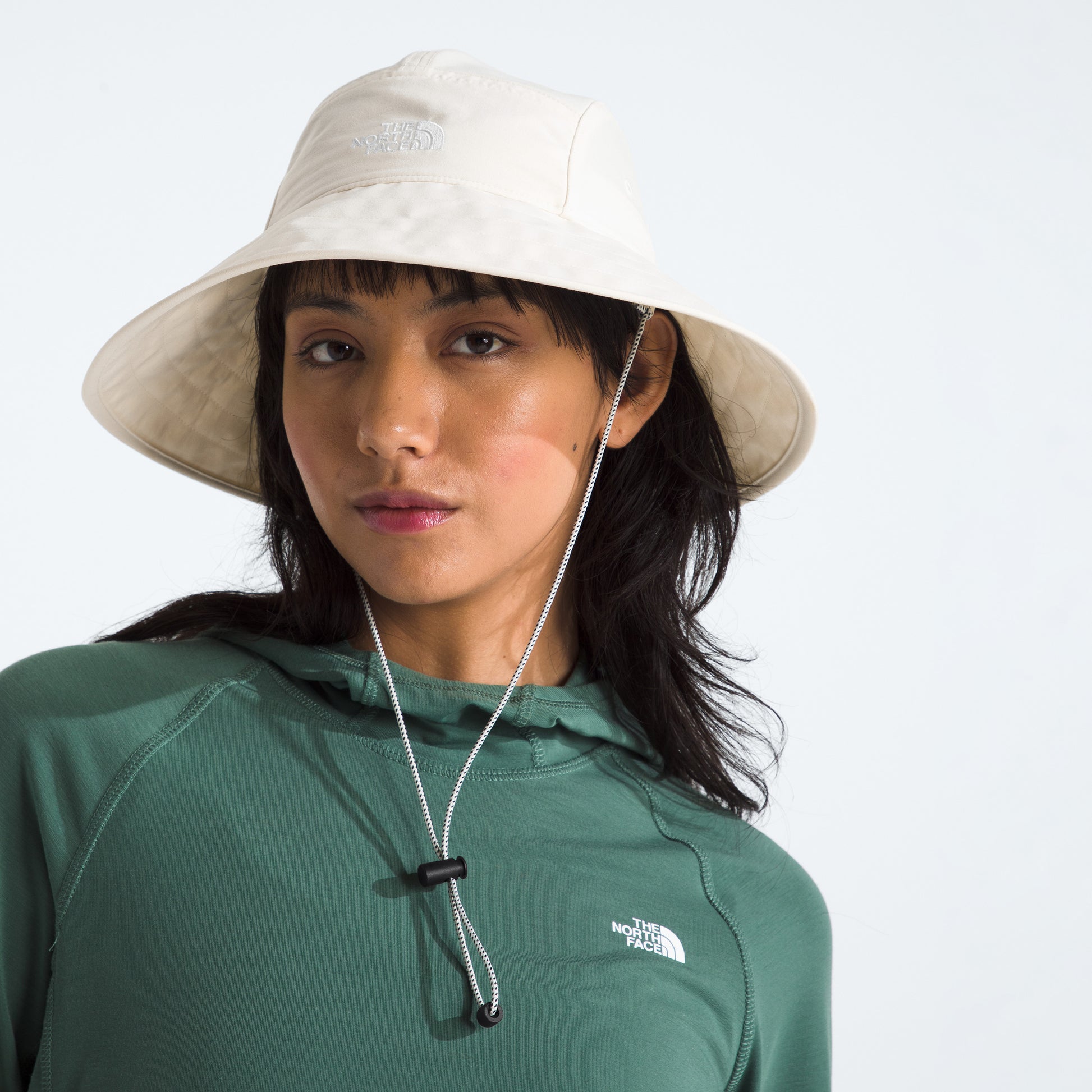 A person in a green top and a cream 'The North Face W Class V Brimmer' Hat with a chin strap, offering sun protection, gazes confidently at the camera.
