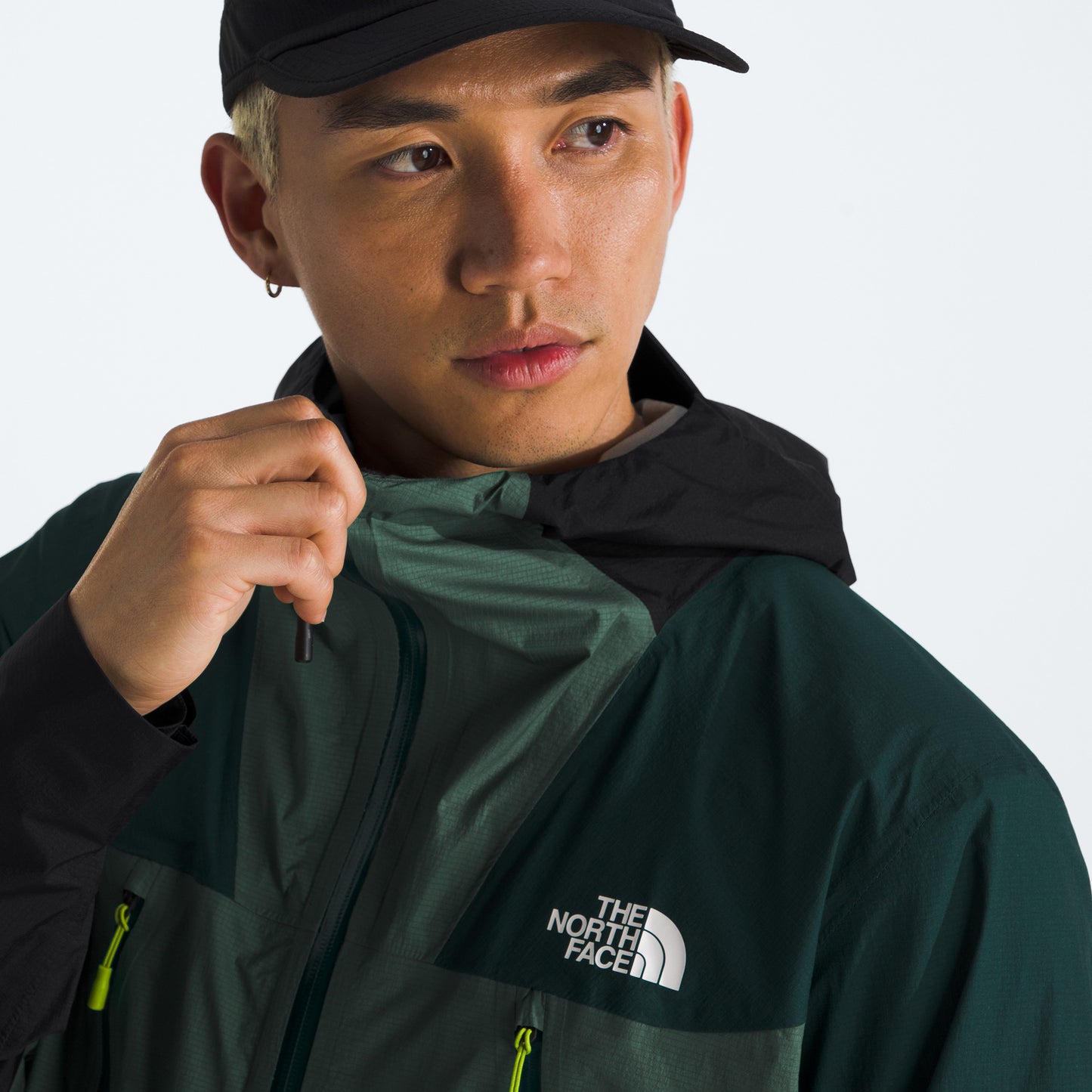 A person in a green and black M Signal 2.5L DryVent jacket by The North Face, wearing a black cap, adjusts the zipper while gazing to the side.