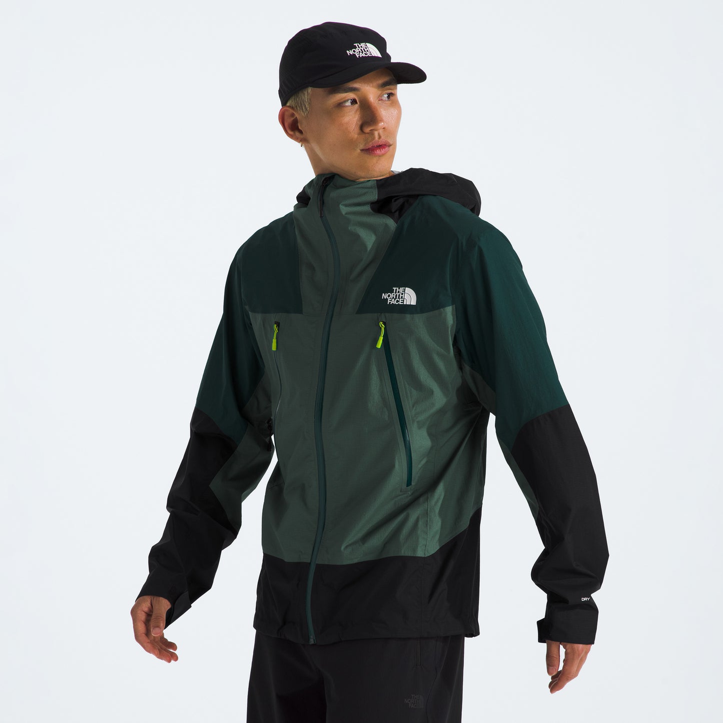 A person wearing a green and black M Signal 2.5L DryVent jacket and a black cap, both featuring The North Face's white logo, standing against a plain background.