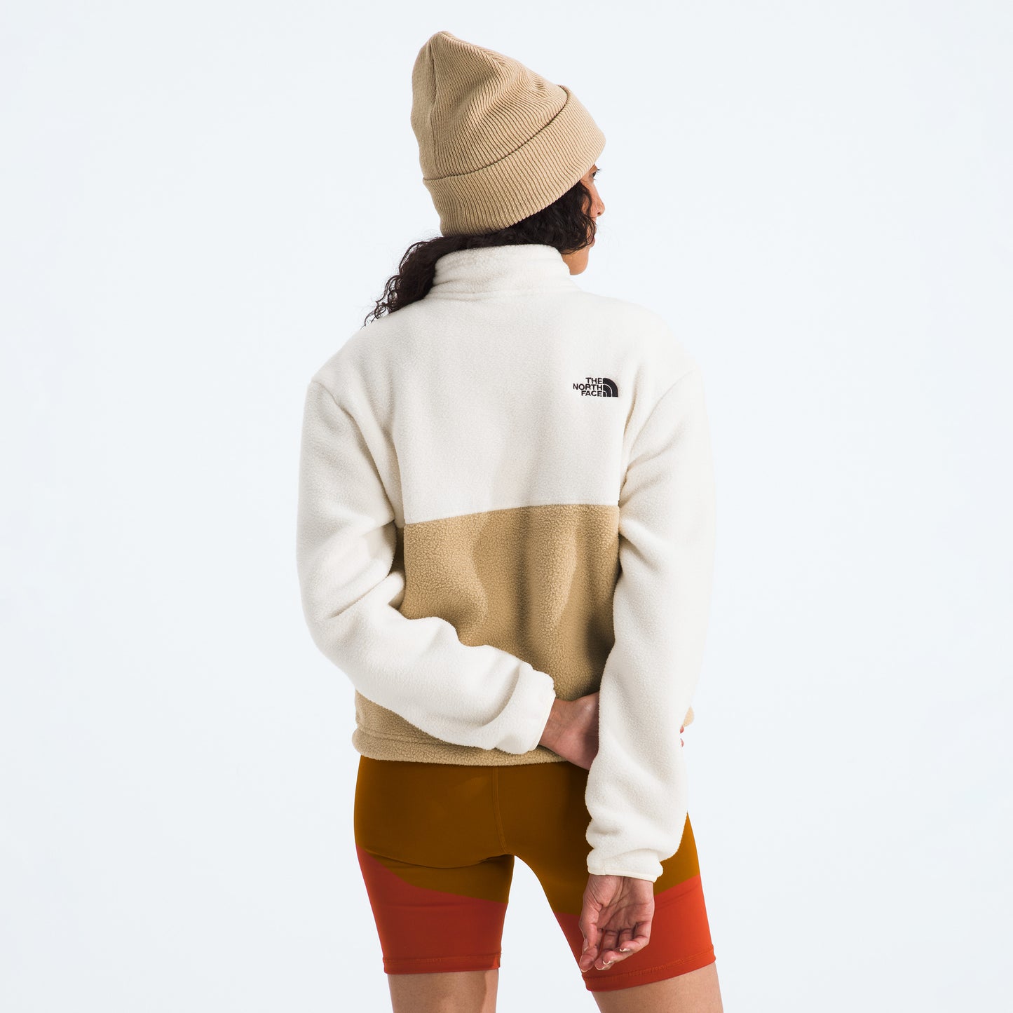 A person in The North Face's W Yumiori 1/4 Zip recycled fleece and a beanie, facing away with one arm behind their back, wearing red-orange shorts.