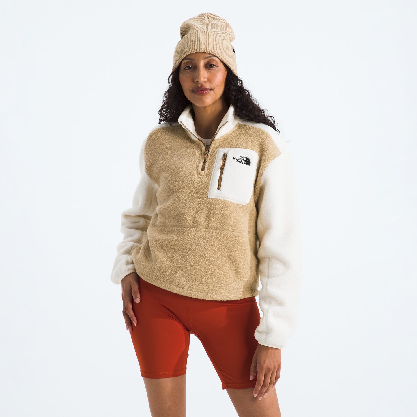 A person wearing The North Face W Yumiori 1/4 Zip in beige fleece, complemented by red shorts and a matching beige beanie, stands against a plain white background.