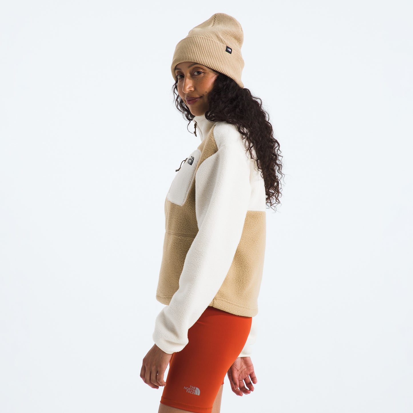 A person wears The North Face's sustainably made W Yumiori 1/4 Zip in beige and white, paired with a beige beanie and orange shorts, standing against a plain background, glancing over their shoulder.