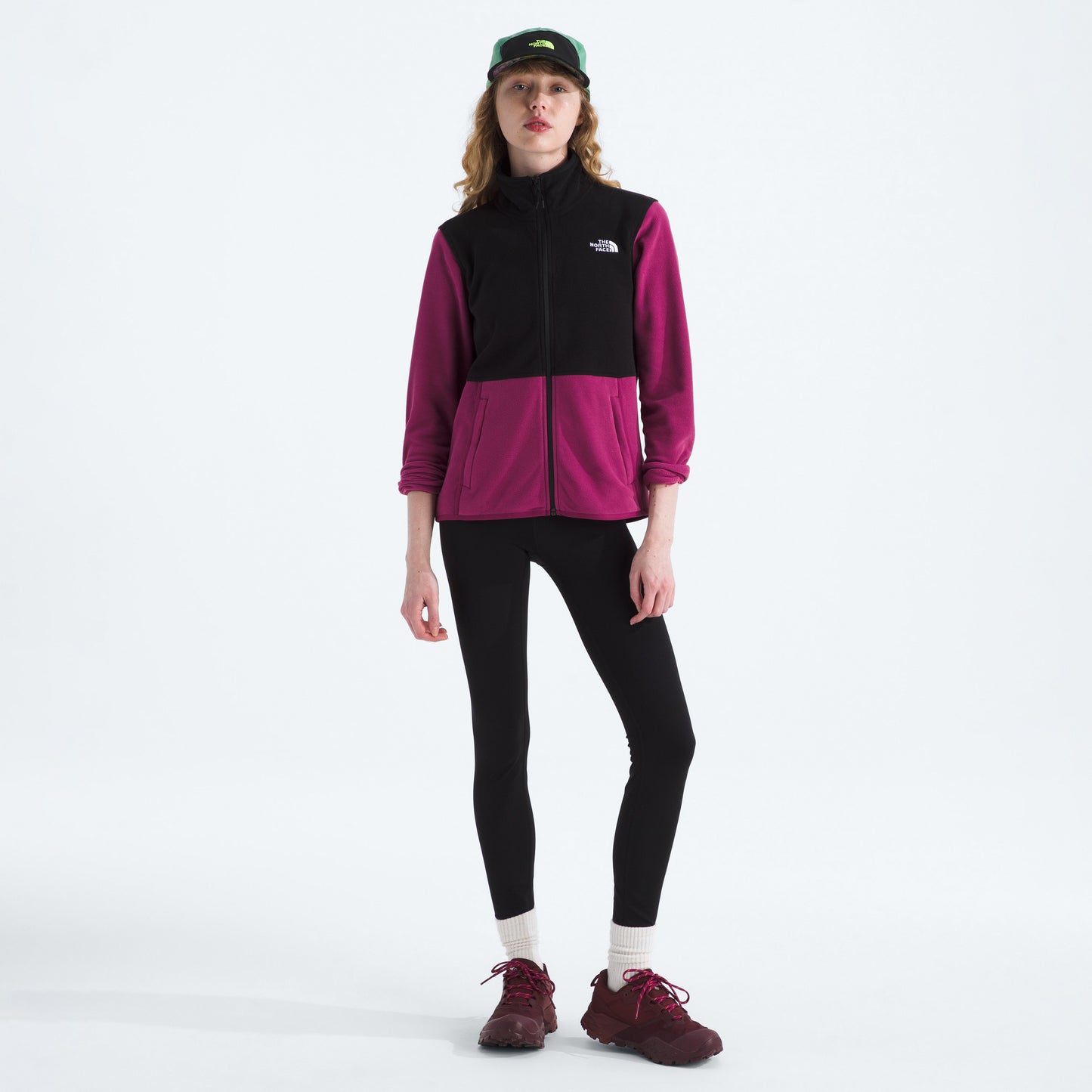 A person in a black and magenta W Glacier Fleece Jacket by The North Face, made from recycled fabric, pairs it with black leggings, maroon shoes, and a green cap against a white background.