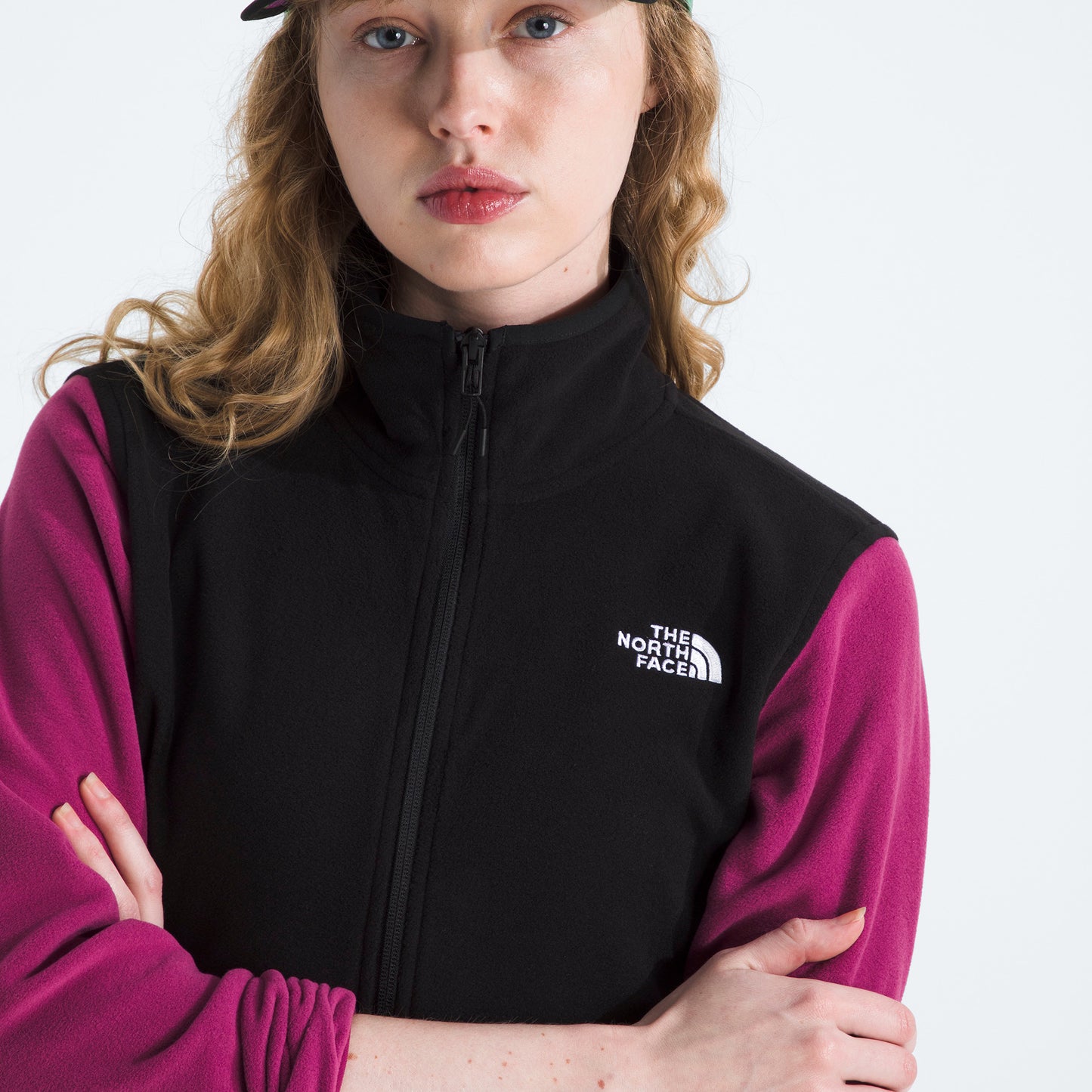 A person with wavy hair, arms crossed, wearing The North Face’s black and magenta W Glacier Fleece Jacket made from recycled fabric, paired with a baseball cap.