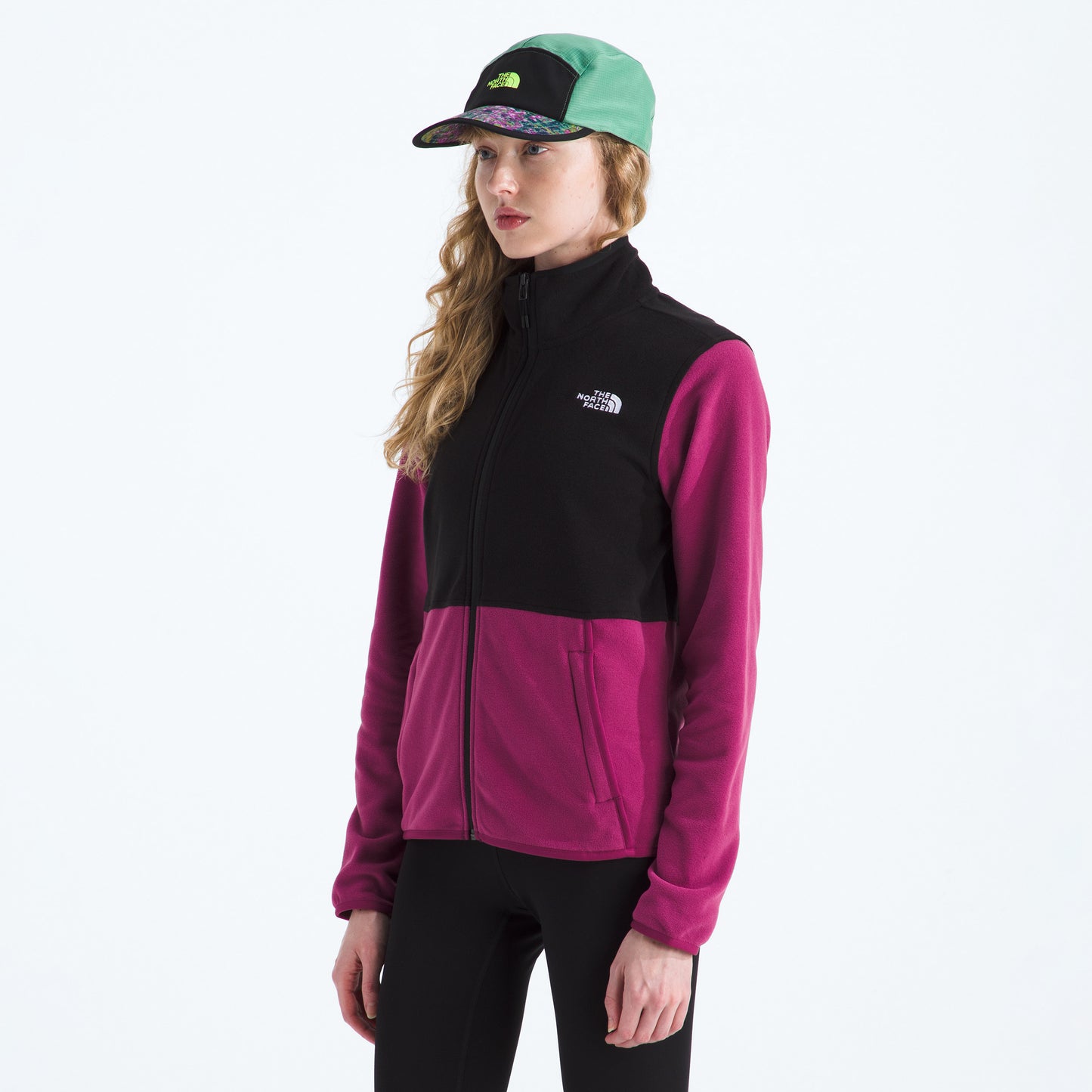 A person wearing The North Face W Glacier Fleece Jacket, featuring a unique black and pink design, pairs it with black leggings and a green-and-black cap against a white background. This lightweight fleece combines eco-friendly recycled fabric with style and comfort.