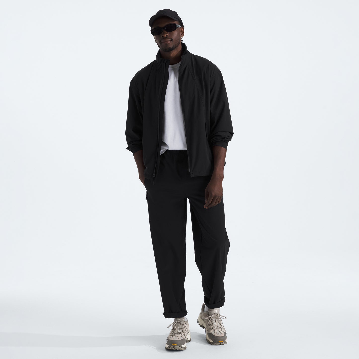 A person stands against a plain white background wearing a black jacket, white shirt, The North Face sneakers, sunglasses, a cap, and the M North Dome Wind Pant by The North Face.