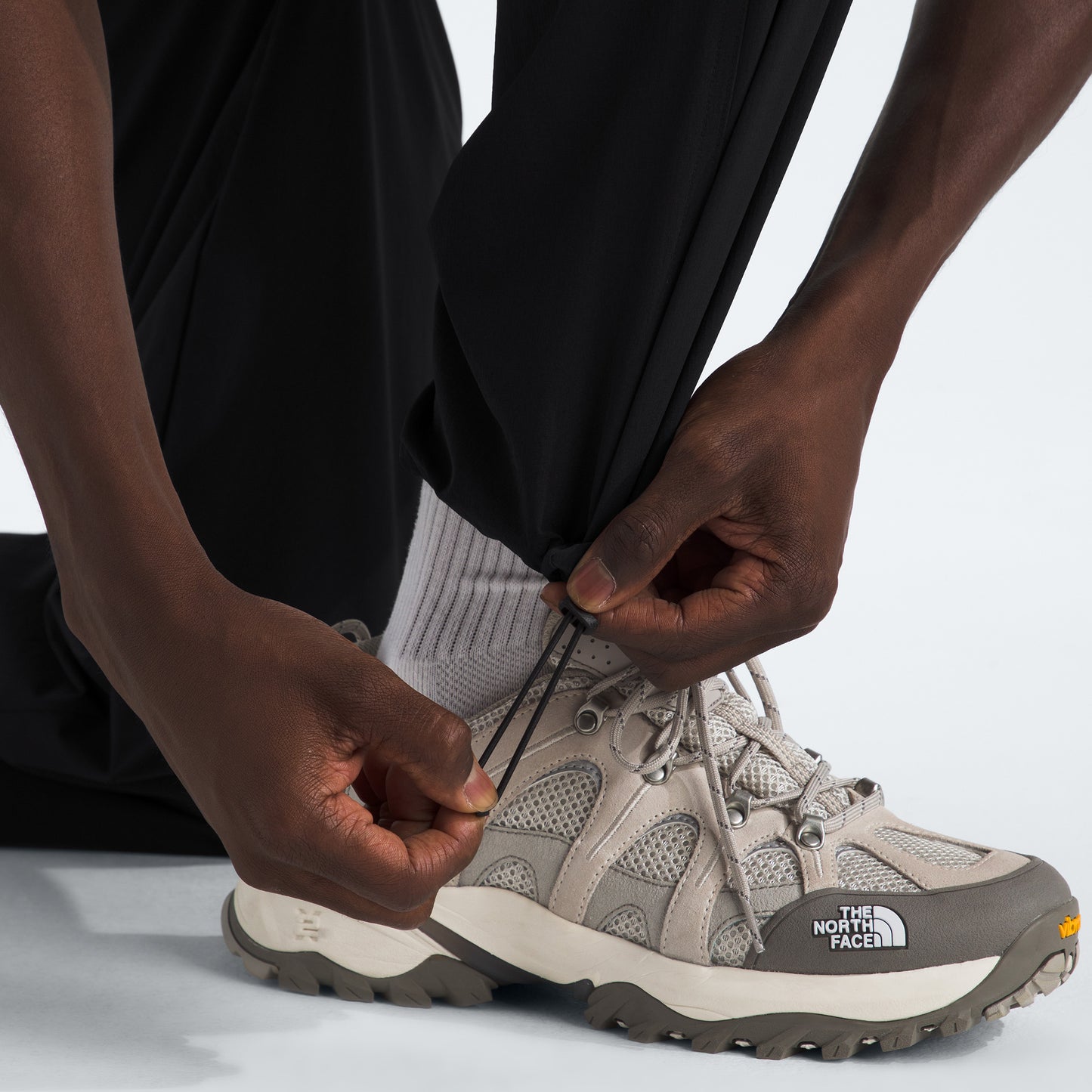 A person adjusts the drawstring hem of The North Face M North Dome Wind Pant, paired with light gray The North Face sneakers and white socks.