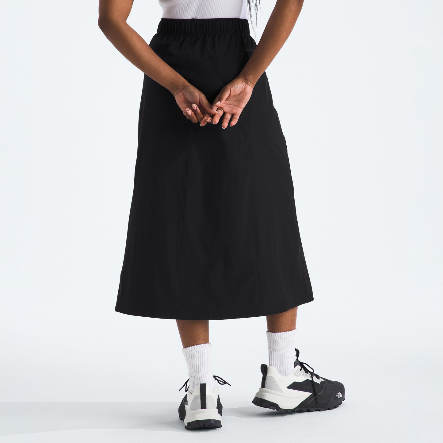 A person stands with their back to the camera, hands clasped behind, wearing The North Face W North Dome Wind Skirt (black), white socks, and black sneakers.