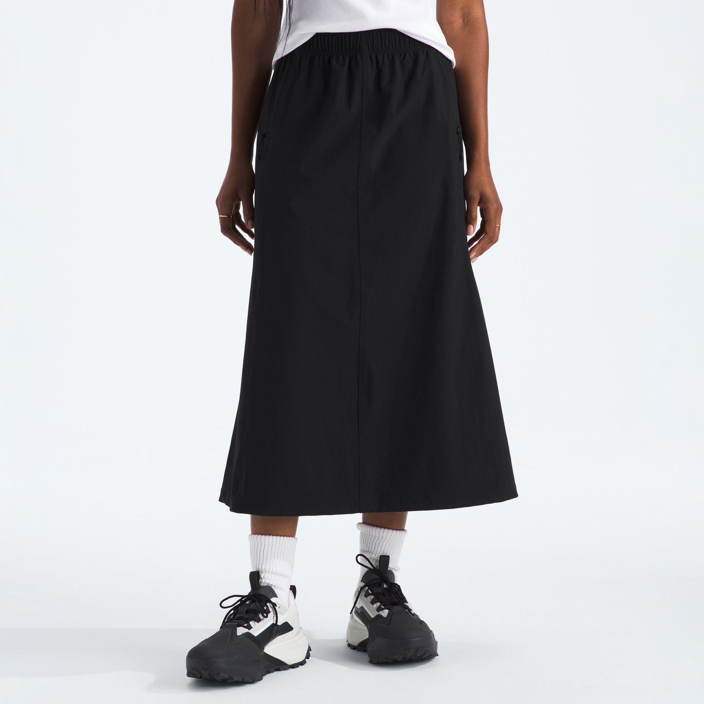 A person dressed in The North Face's W North Dome Wind Skirt stands on a plain background, wearing white socks and black-and-white sneakers.