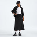 A person is wearing a black cap, jacket, W North Dome Wind Skirt by The North Face, white shirt, and sneakers, standing against a plain background.
