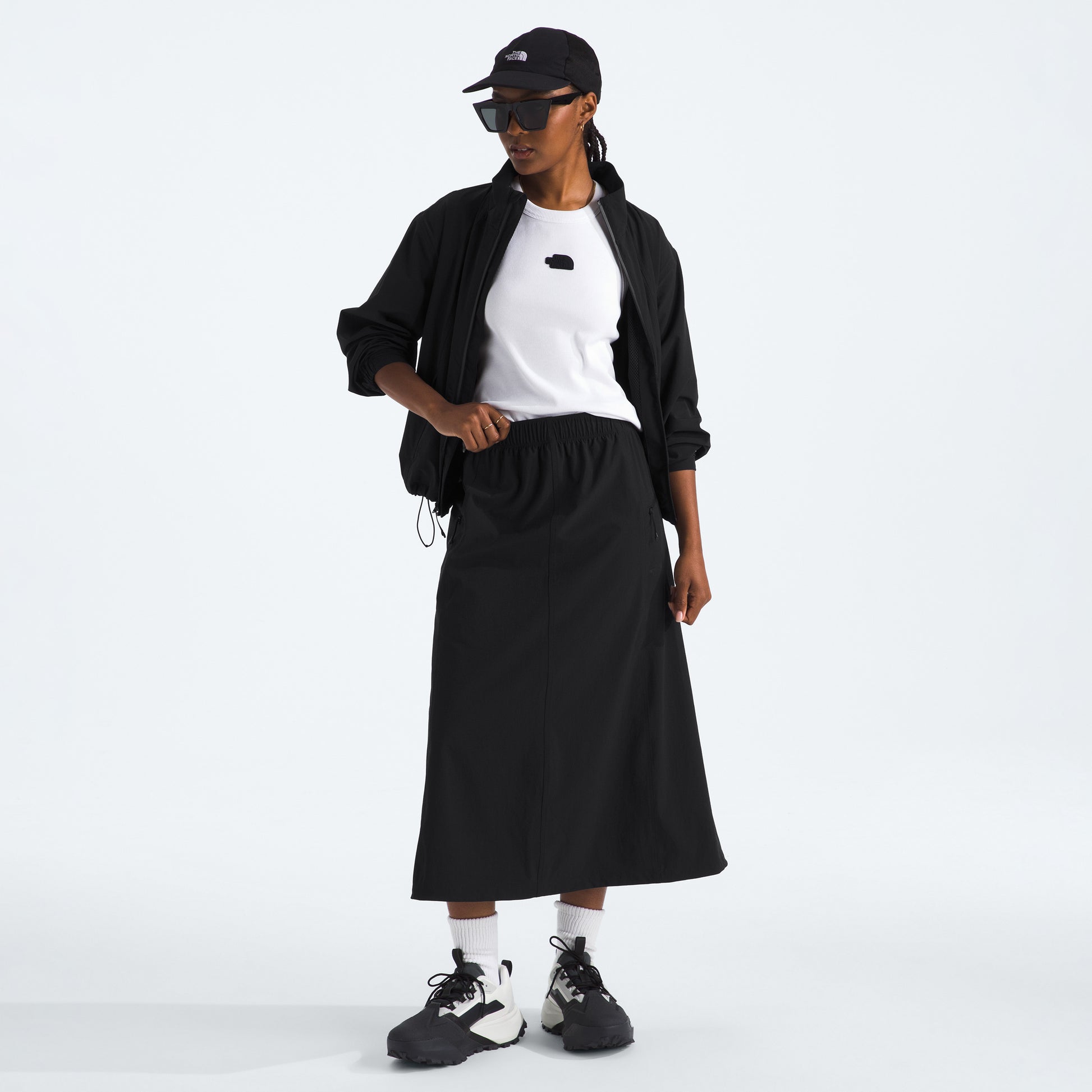 A person is wearing a black cap, jacket, W North Dome Wind Skirt by The North Face, white shirt, and sneakers, standing against a plain background.